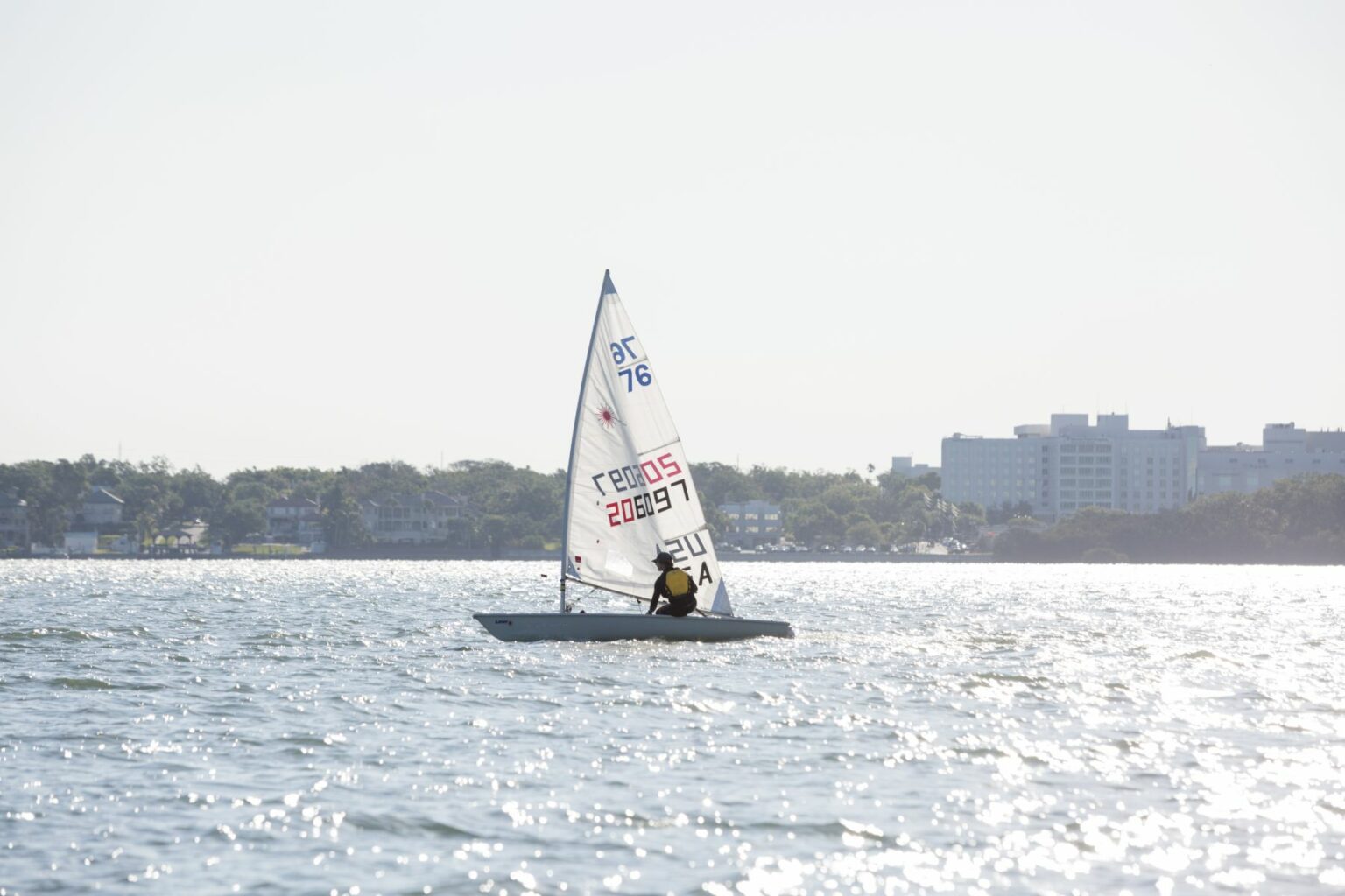 License-free image - A lone sailor navigates a small sailboat on a shimmering, sunlit body of water, with a distant shoreline and cityscape visible in the background. The sail is marked with various numbers and letters. Sailing while wearing a properly fitting life vest in the Sand Key inlet in Clearwater Beach.