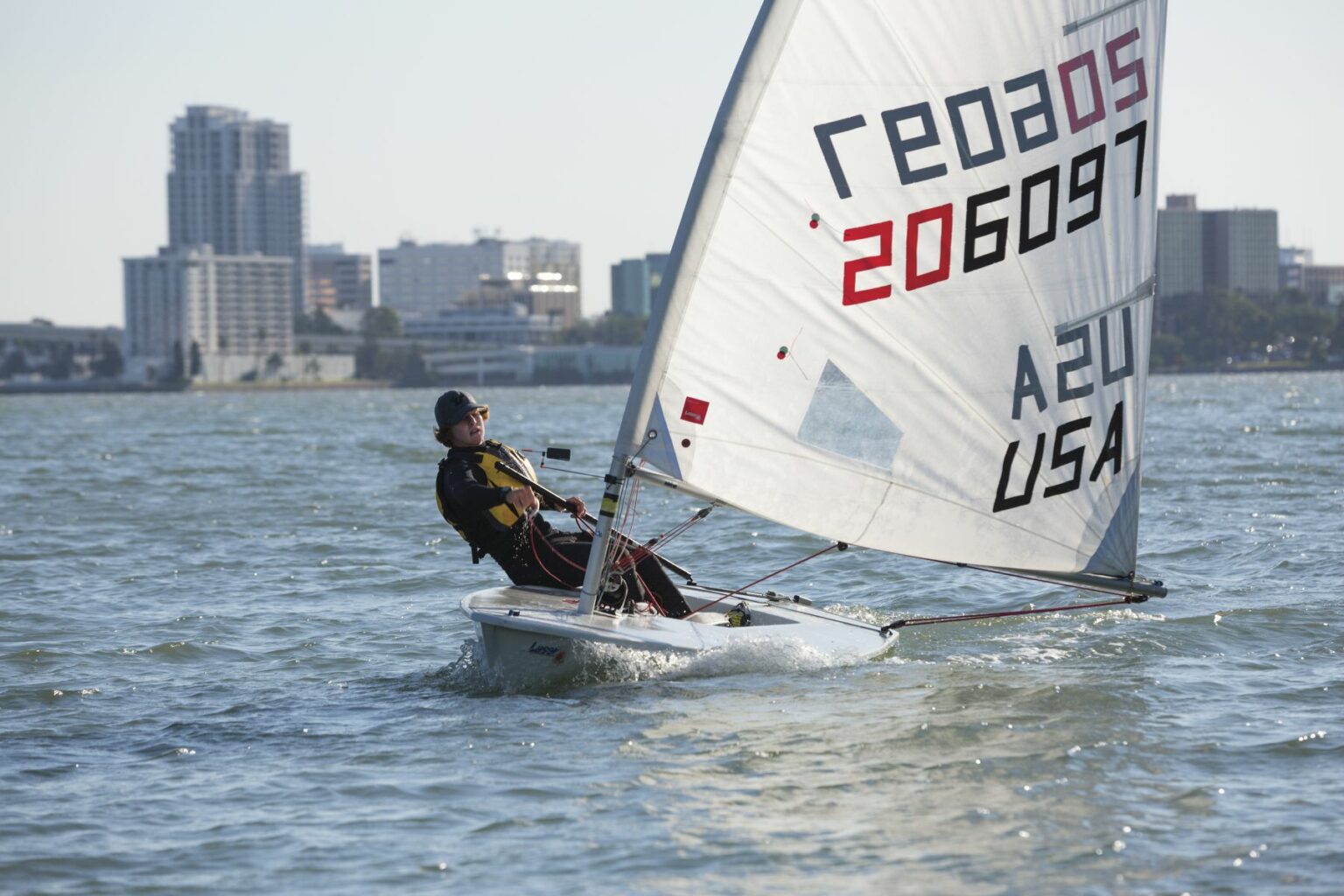 License-free image A person is sailing a small boat on a body of water with a cityscape in the background. The sail displays the numbers &quot;206097&quot; along with &quot;A24&quot; and &quot;USA.&quot; The sky is clear and sunny. Sailing while wearing a properly fitting life vest in the Sand Key inlet in Clearwater Beach.