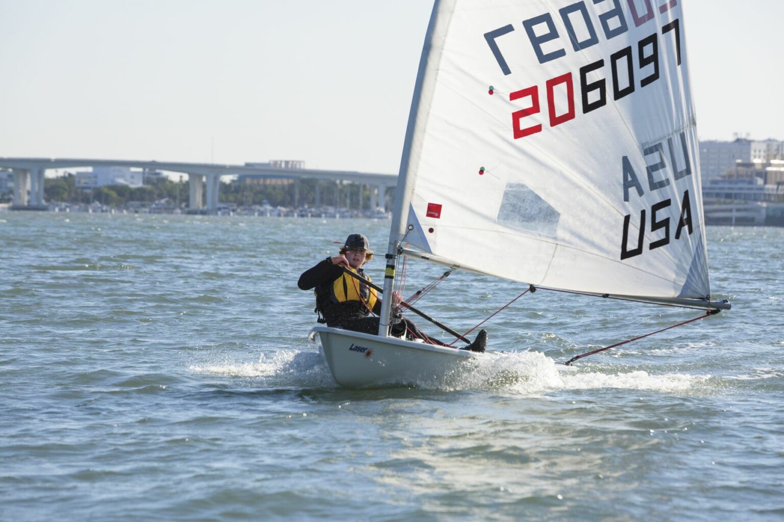 License-free image A sailor navigates a small sailboat on calm waters with a cityscape and bridge in the background. The sail has markings &quot;206097&quot; and &quot;USA.&quot; The sailor is wearing a hat and life jacket, holding onto the sail and the tiller. The water shows slight ripples. Sailing while wearing a properly fitting life vest in the Sand Key inlet in Clearwater Beach.