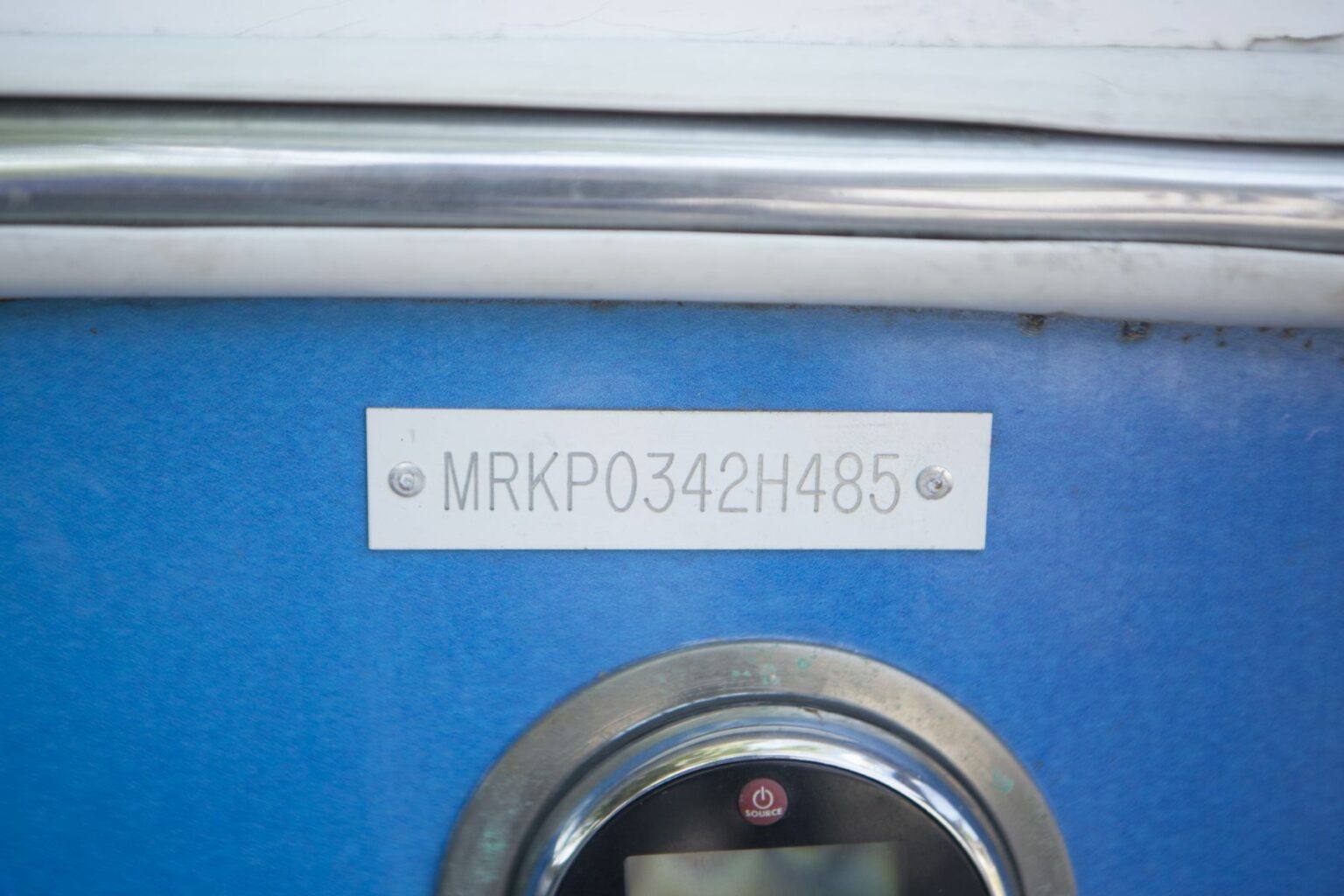 License-free image - Close-up of a metal plate Hull Identification Number attached to a blue boat hull surface with the text &quot;MRKP0342H485&quot; engraved on it. Below the plate is a circular device with a small screen and a button marked by a red emblem. Hull identification number on a watercraft.