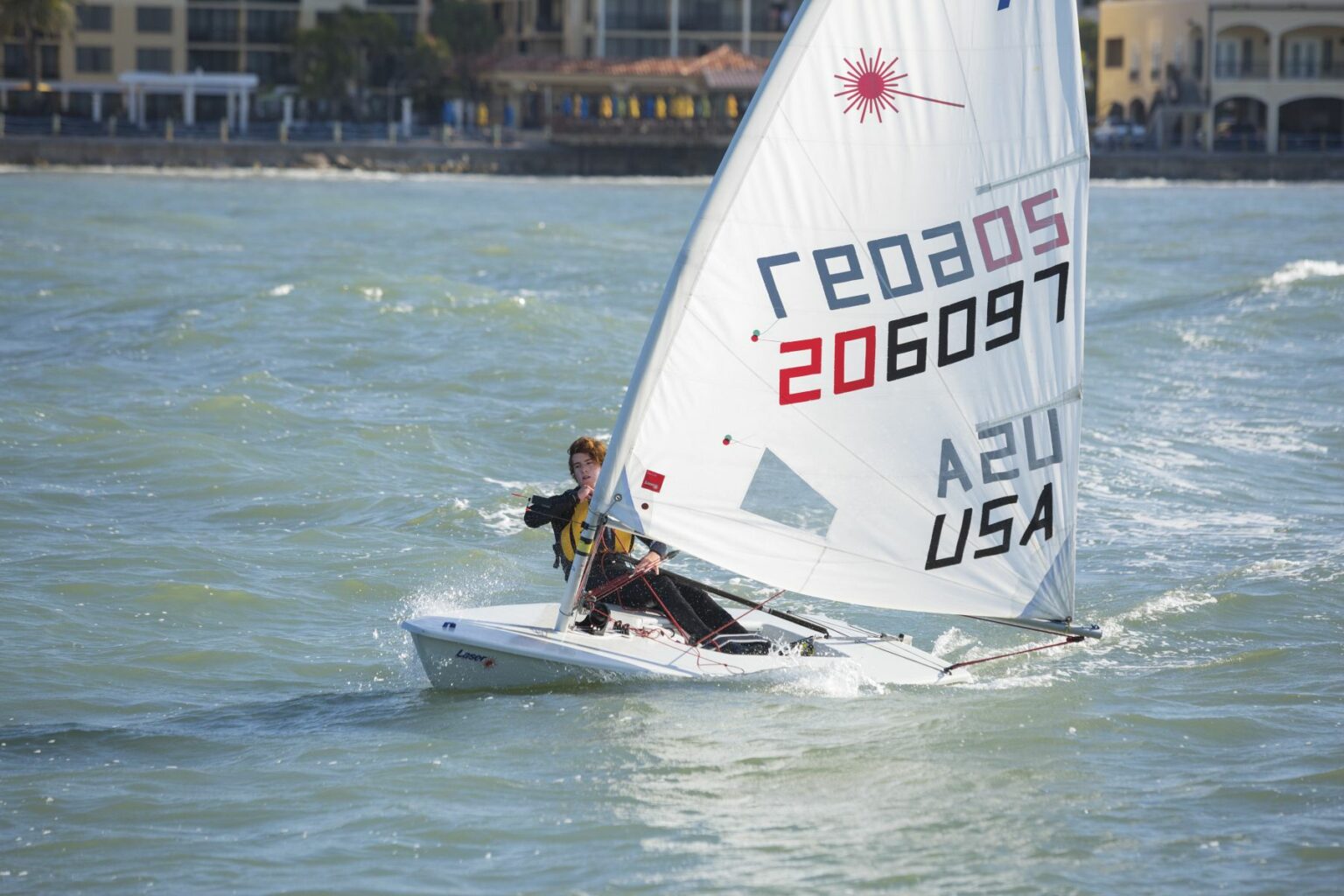 License-free image A person is sailing a small boat with a white sail on a body of water. The sail has various numbers and letters printed, including &quot;206097&quot; and &quot;USA.&quot; Buildings are visible in the background along the shoreline. The weather appears to be clear and sunny. Sailing while wearing a properly fitting life vest in the Sand Key inlet in Clearwater Beach.