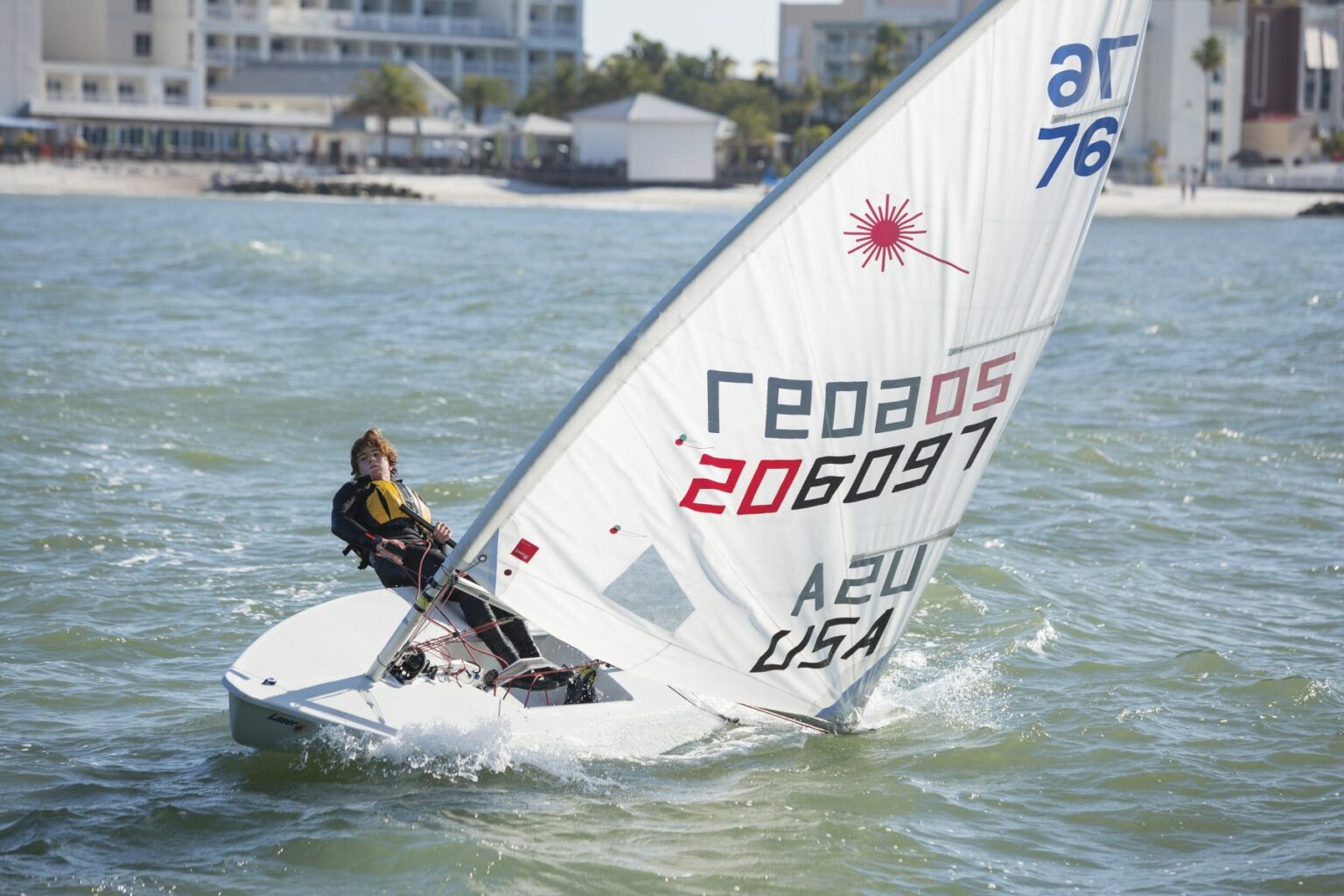 License-free image A person wearing a life jacket and harness is sailing a small boat with a white sail on choppy water. The sail has numbers and a red logo. Buildings and trees are visible in the background near the shoreline. Sailing while wearing a properly fitting life vest in the Sand Key inlet in Clearwater Beach.