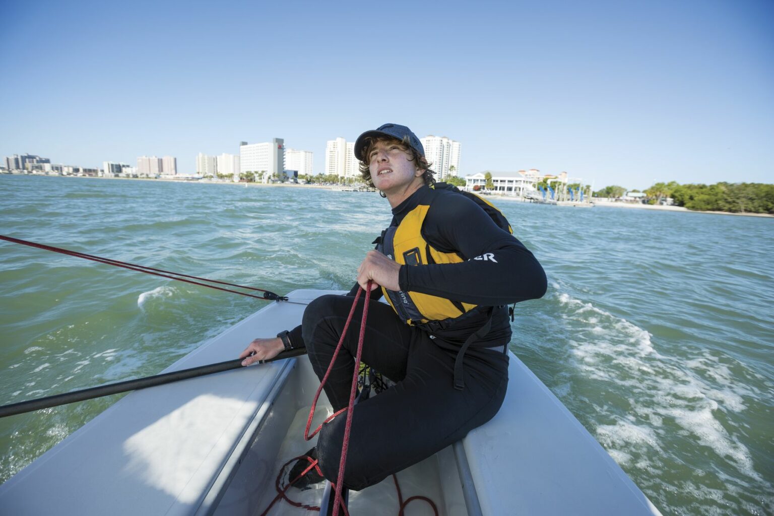 License-free image A person in a black wetsuit and yellow life vest sits in a small boat holding a red rope, with a city skyline of tall buildings in the background and green water surrounding them. They appear focused, looking towards the horizon, under a clear blue sky. Sailing while wearing a properly fitting life vest in the Sand Key inlet in Clearwater Beach.