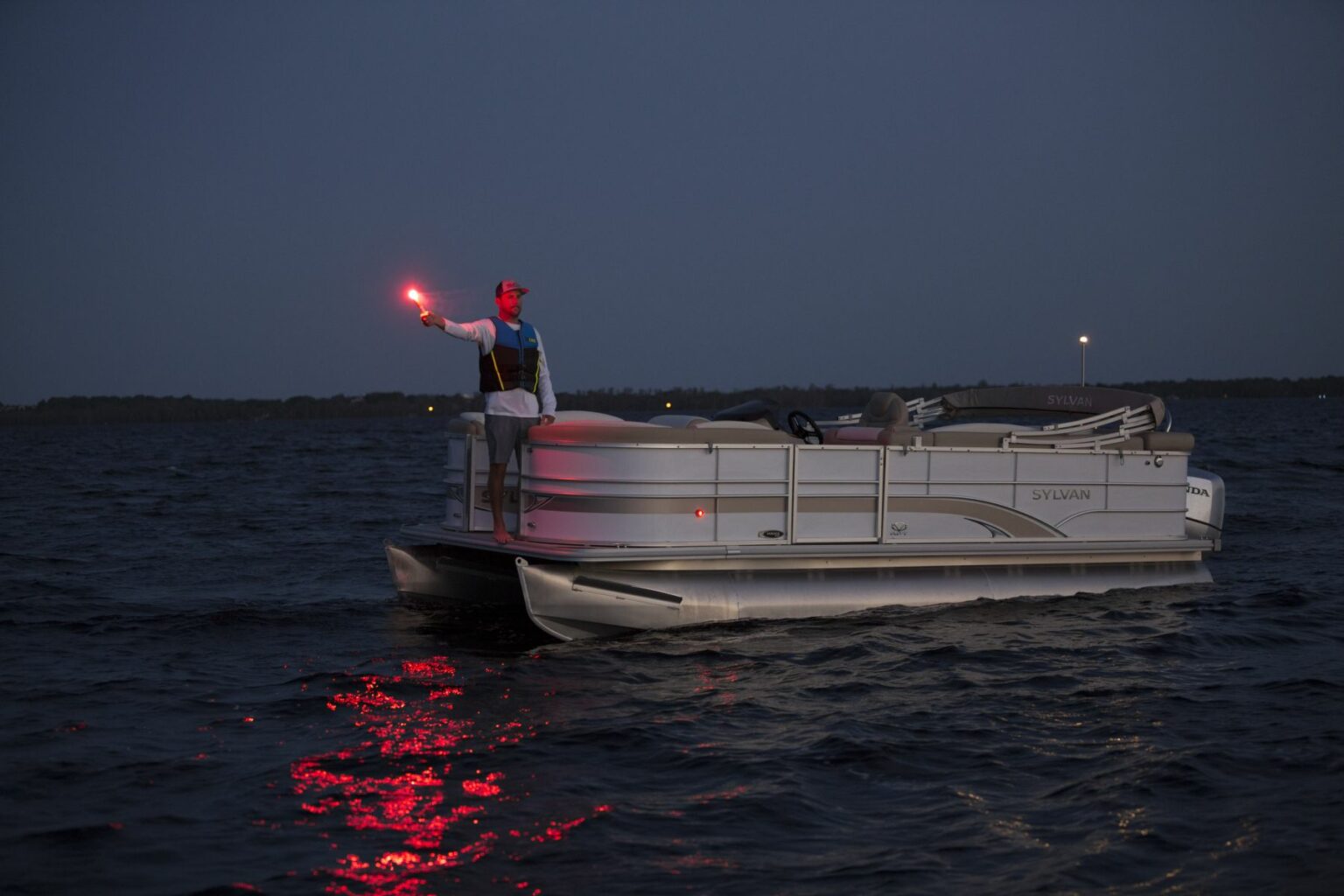 License-free image -  Person standing on a pontoon boat at dusk, holding a red flare aloft in their right hand. The water around the boat reflects the red light from the flare. The person is wearing a hat and a life vest. In the background, the horizon is visible under a darkening sky.