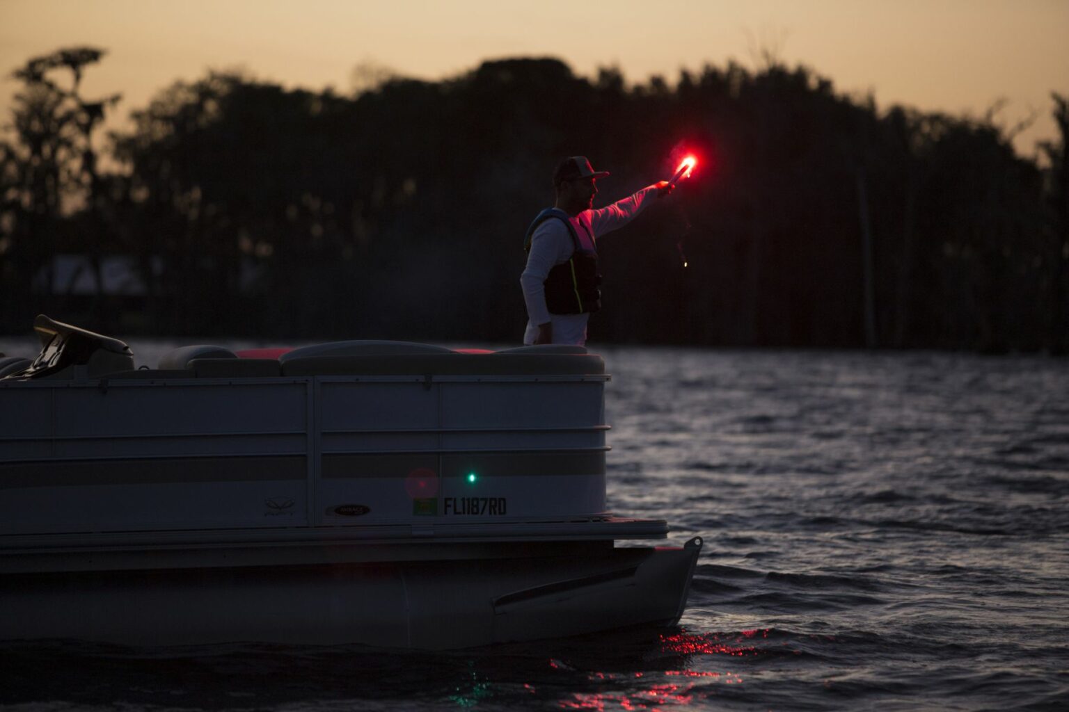 License-free image - A person stands on the edge of a boat at dusk, holding a red flare. The silhouette of trees is visible in the background, contrasting against the darkening sky. The water around the boat is calm with slight ripples.