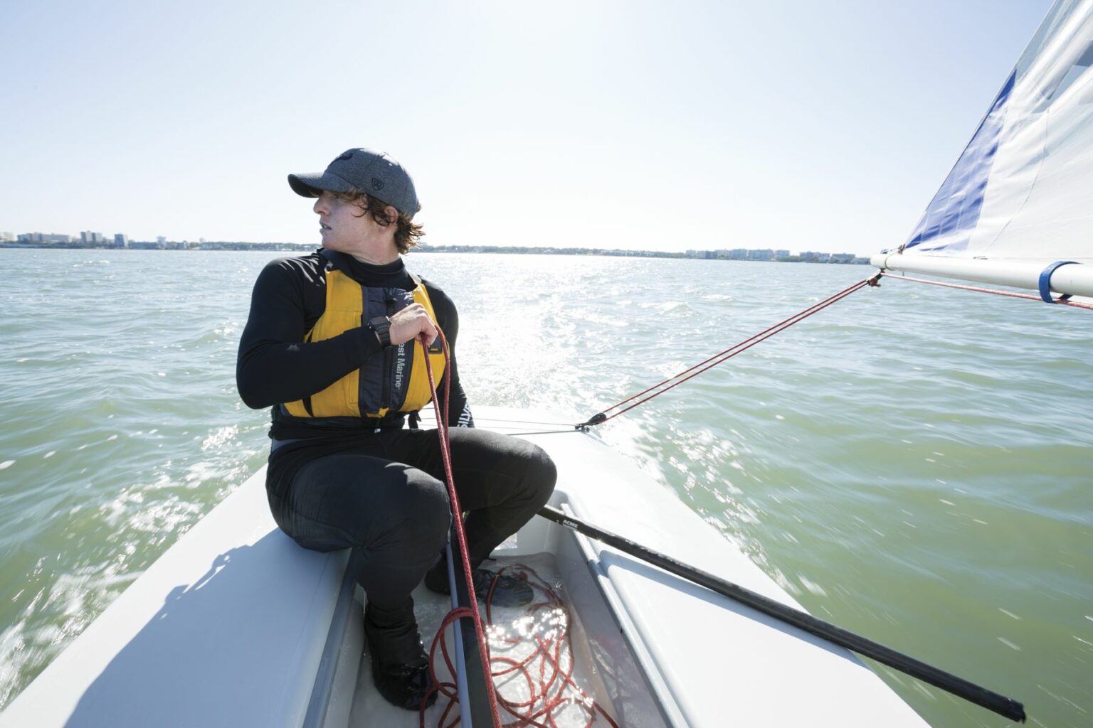 License-free image - A person in a yellow life vest and black clothing is sailing a small boat on a sunny day. They are looking to the left while holding a red rope. The water is calm and a distant shoreline is visible in the background. Sailing while wearing a properly fitting life vest in the Sand Key inlet in Clearwater Beach.