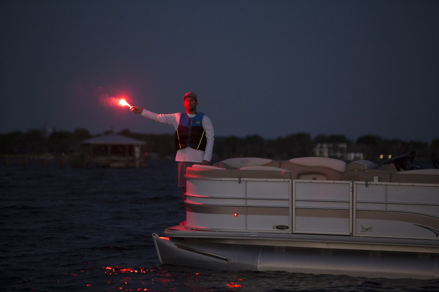 Royalty-free image - A person stands at the edge of a boat, holding up a lit red flare against the night sky. They are wearing a white shirt and a life jacket. The water and distant shoreline with buildings are visible in the background.