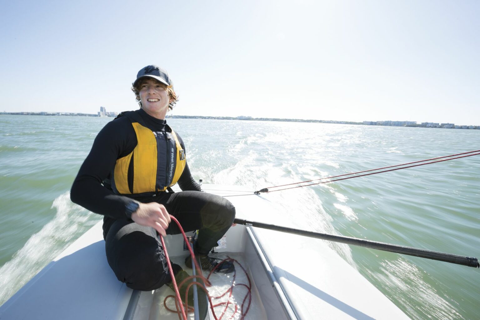 Royalty-free image - A person wearing a yellow and black life jacket and a black cap is sailing on a small boat in open waters on a sunny day. The individual is smiling and holding a rope, with a cityscape visible in the distant background. Sailing while wearing a properly fitting life vest in the Sand Key inlet in Clearwater Beach.