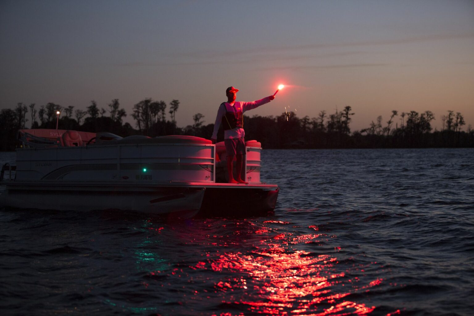 License-free image - A person stands on a boat at dusk holding a red flare aloft, illuminating the surrounding water with a red glow. Trees and a dimly lit sky are visible in the background. The boat is reflected in the water below, creating a calming and dramatic scene.