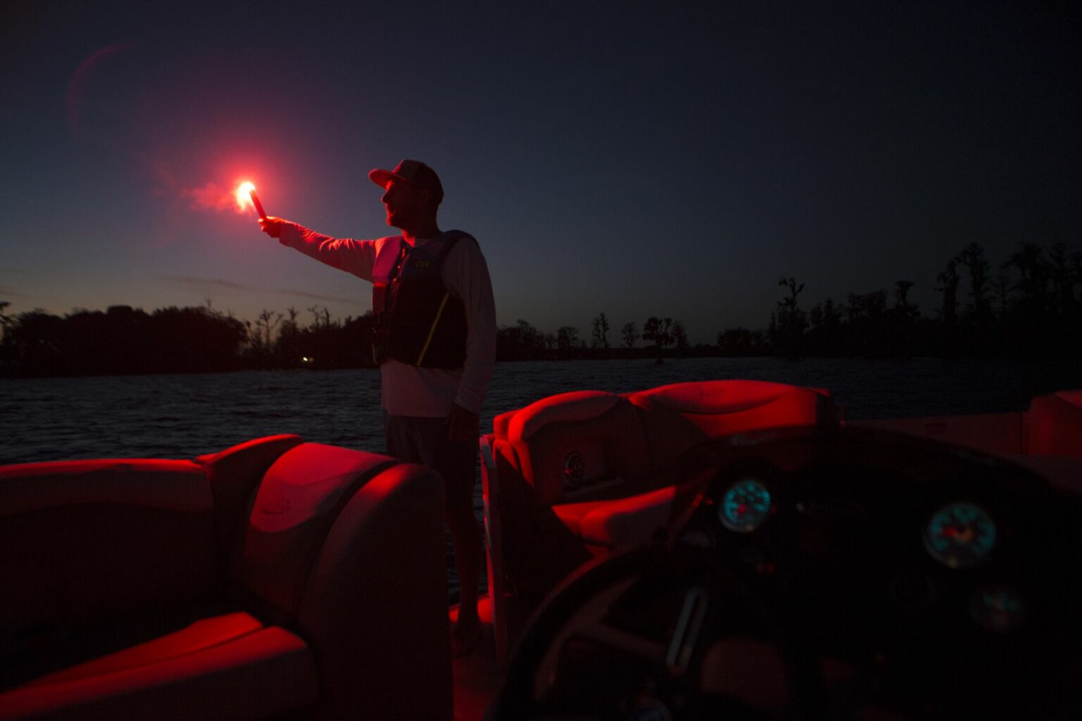 License-free image - A person standing on a boat at dusk or nighttime, holding up a red flare. The red light illuminates their silhouette and the interior of the boat. Trees and a dark sky are visible in the background. The boat&#039;s dashboard with illuminated gauges is also visible.