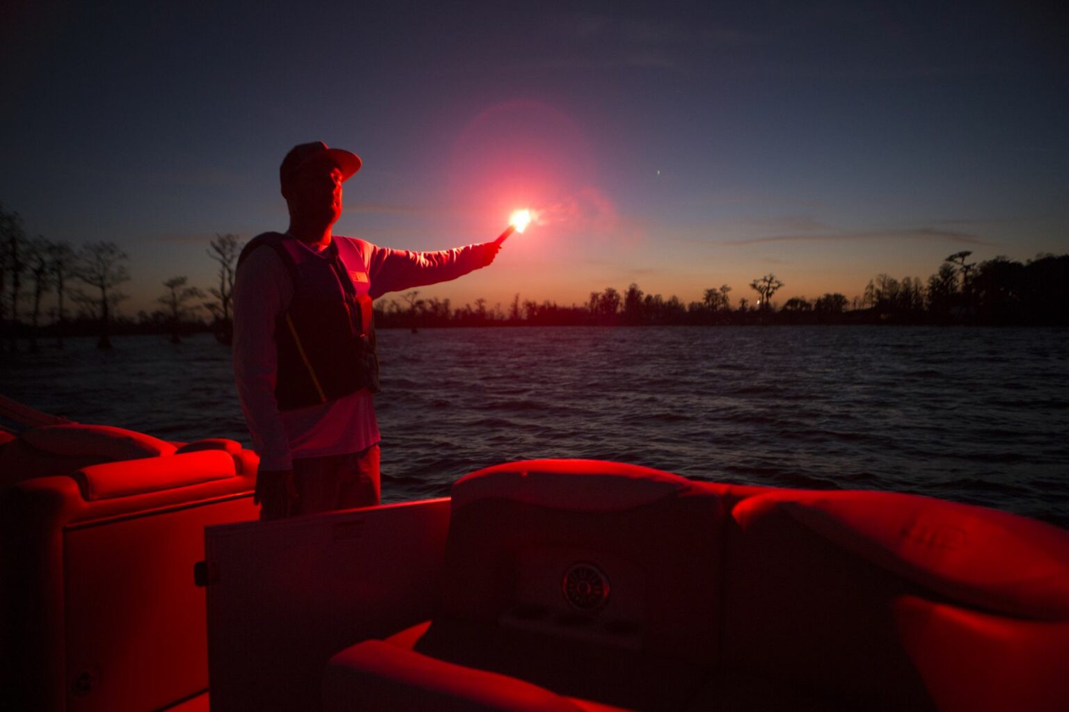 License-free image A person stands on a boat at dusk, holding a red flare that illuminates the scene. The sky is dark with a fading sunset, and the water around the boat appears calm. The silhouette of the person contrasts with the bright light of the flare.