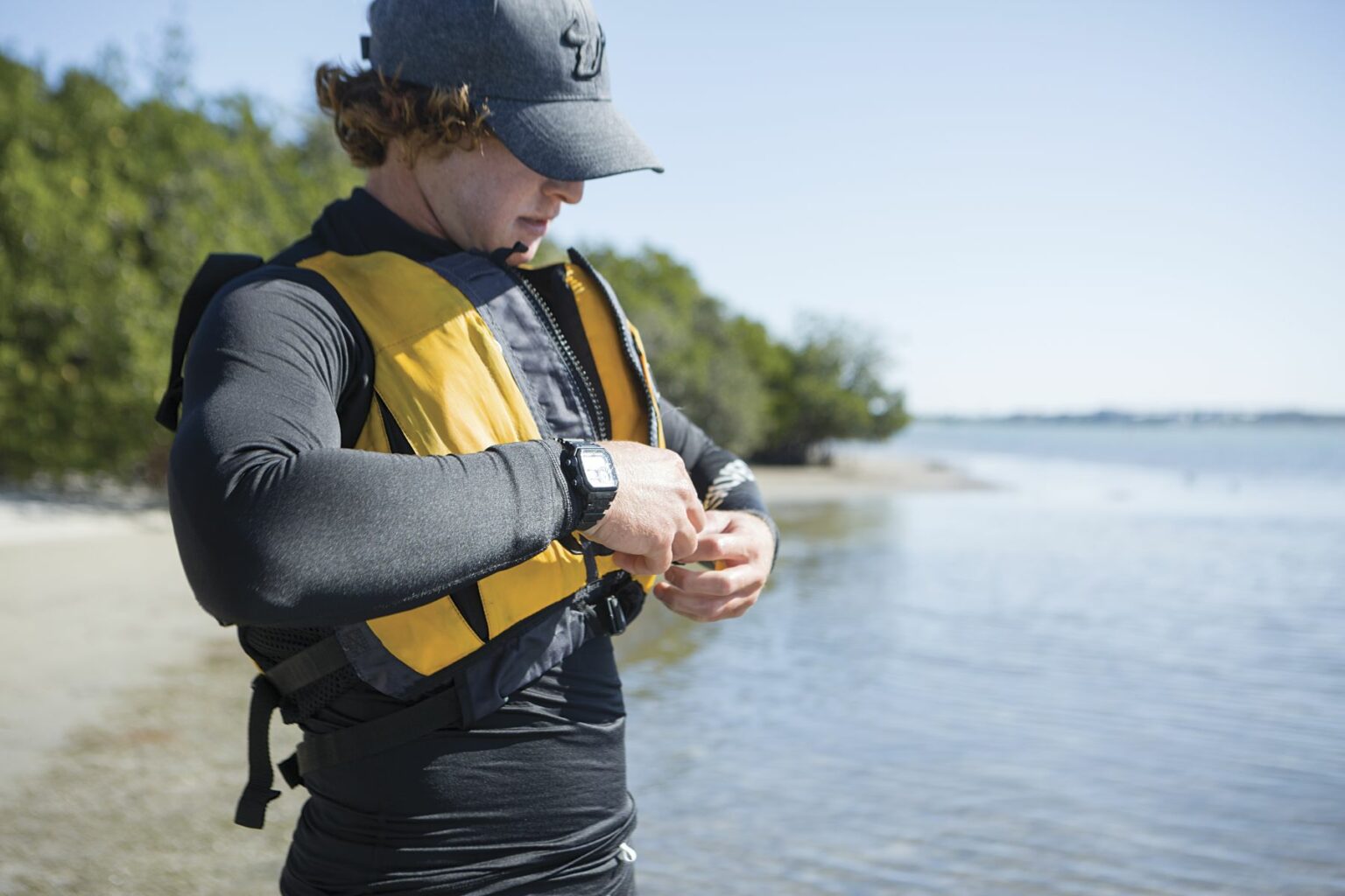 License-free image - A person wearing a grey cap and black long-sleeve shirt adjusts the straps of their yellow life jacket by the water. They stand on a sandy beach with greenery in the background and calm water stretching to the horizon under a clear blue sky. Putting on a properly fitting life vest before sailing in the Sand Key inlet in Clearwater Beach.