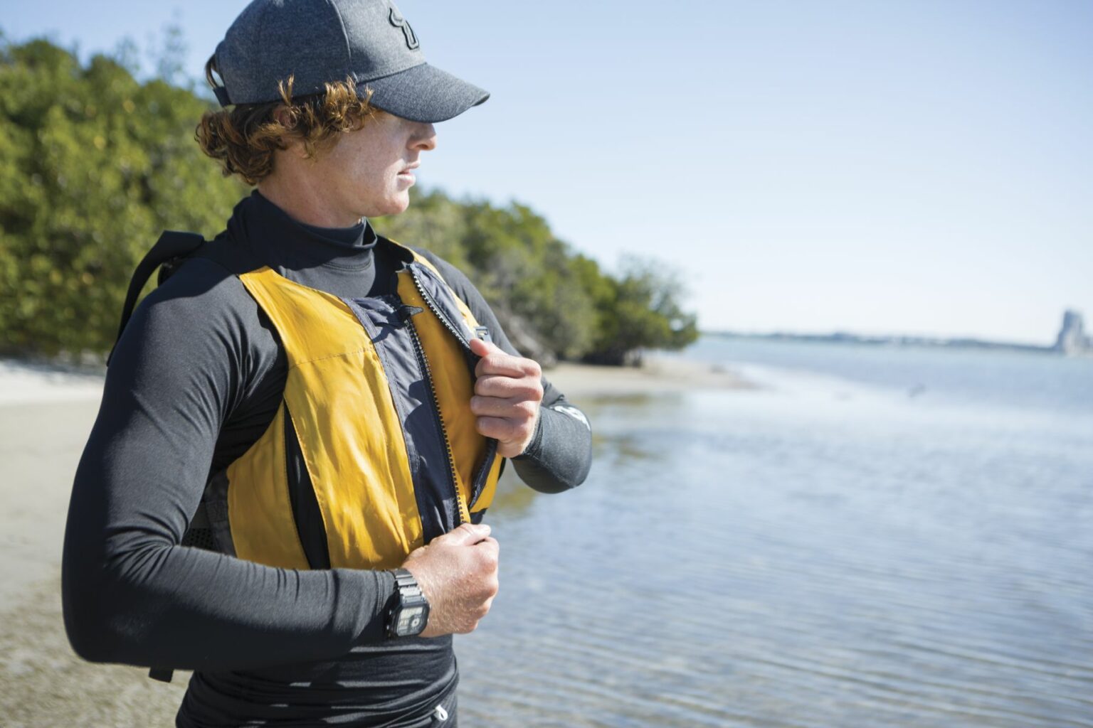 License-free image A person wearing a gray cap and a yellow life jacket stands by the edge of a body of water, zipping up the life jacket. The person appears to be preparing for a water-related activity, with trees and the water visible in the background. Putting on a properly fitting life vest before sailing in the Sand Key inlet in Clearwater Beach.