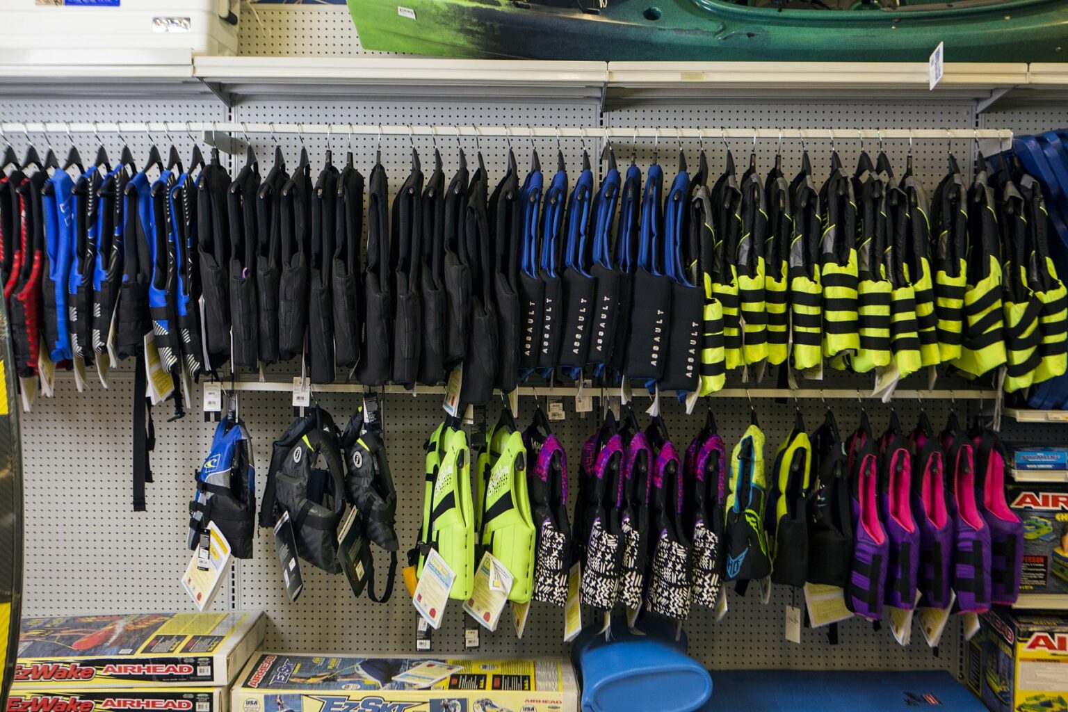 License-free image -  A store display of various life jackets in multiple colors and sizes hangs on a pegboard. Below the life jackets, there are shelves with boxed products and accessories. There is a kayak on the top shelf. Life jackets on display at a retail outlet.