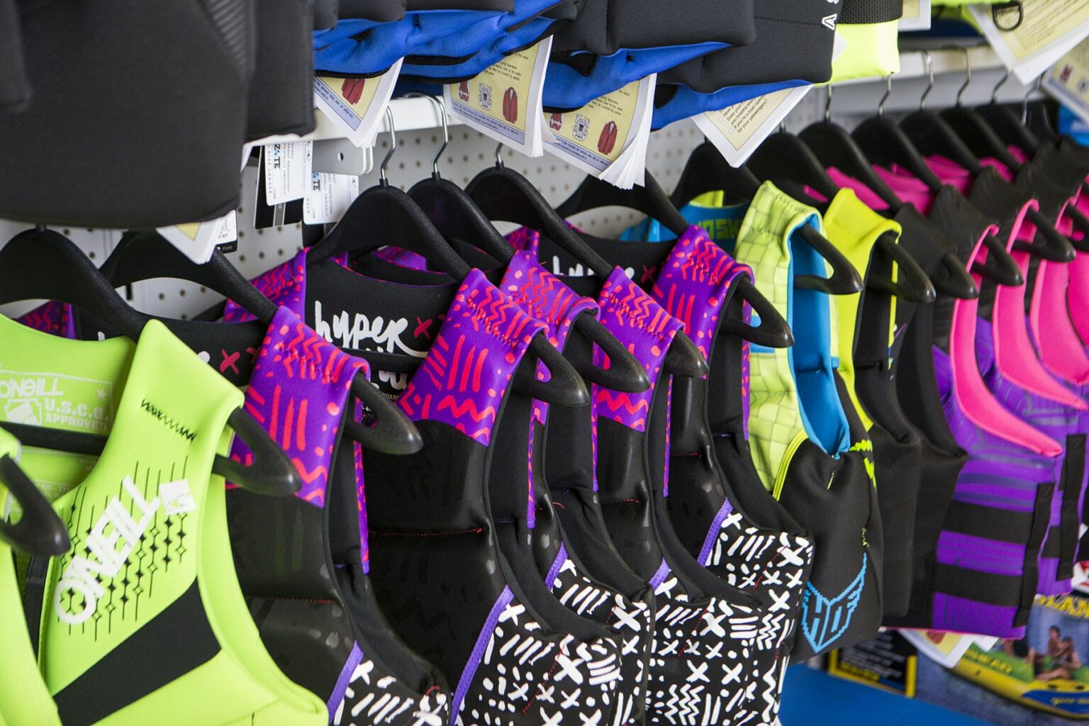 License-free image - A row of colorful life jackets hanging on a retail display, featuring various brands and vibrant designs in shades of yellow, purple, black, and pink. Each life jacket is displayed on a black hanger and has a price tag attached. Life jackets on display at a retail outlet.