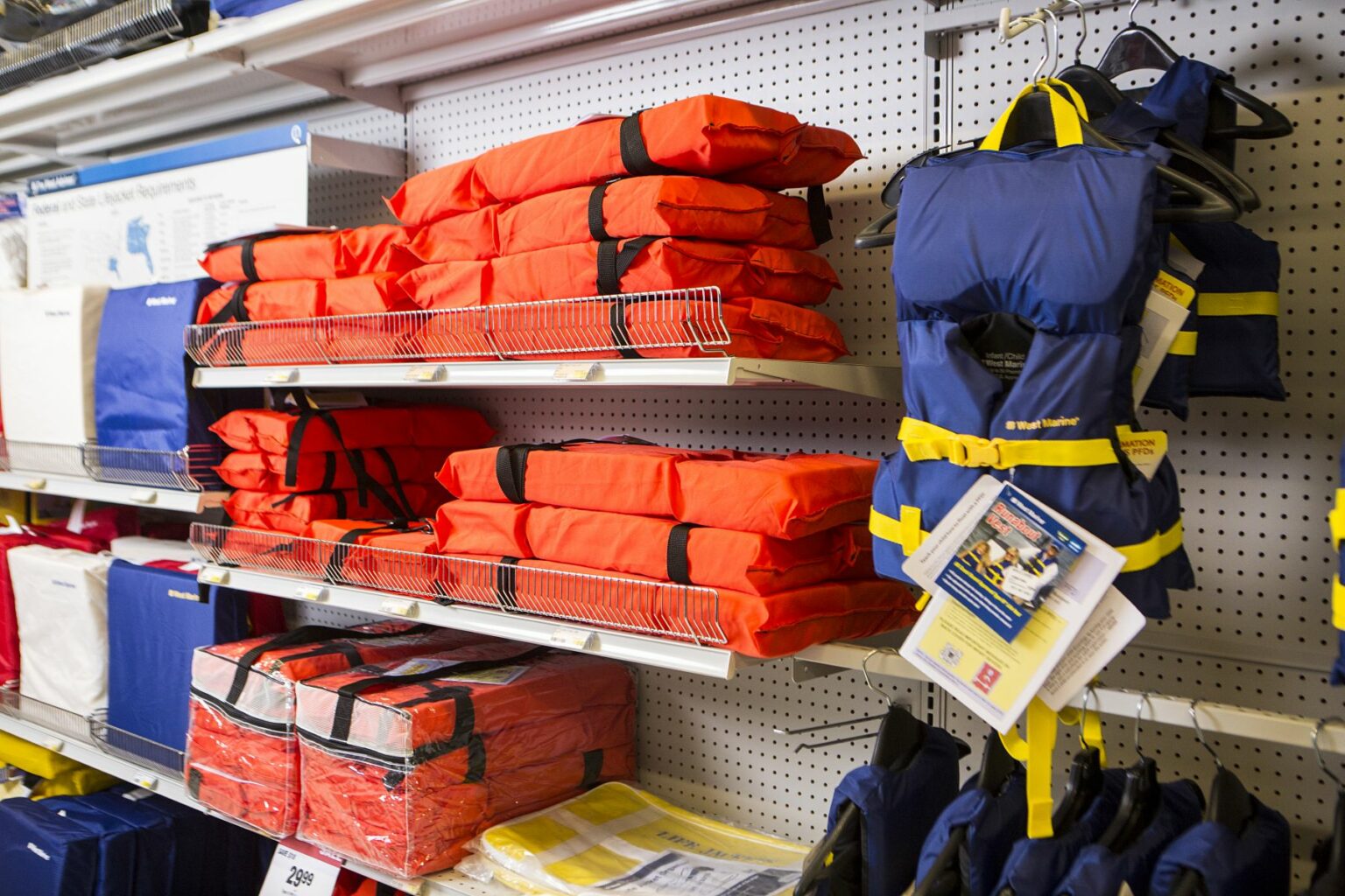 License-free image -  A store display of life jackets. Orange life jackets are neatly stacked on shelves, while blue and yellow life vests are hanging on hooks. The display is well-organized, with additional life-saving gear visible in the background. Life jackets on display at a retail outlet.