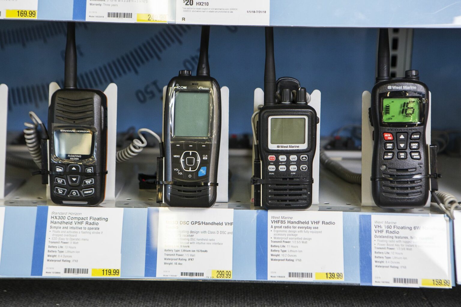 License-free image - A store display shelf with five different handheld VHF radios. Each radio has a description and price tag underneath. The radios vary in design, color, and button layout. The shelf is part of an electronics or marine equipment section. Handheld VHF radios on display at a retail outlet.