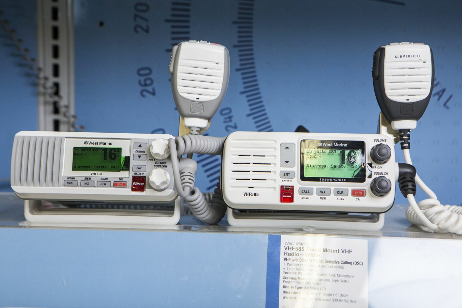 License-free image - Two VHF marine radios with microphones are displayed side by side on a shelf. The radios have digital screens showing frequency information and buttons for various controls. The device on the right also features a larger screen displaying lines of text and an image. VHF radios on display at a retail outlet.