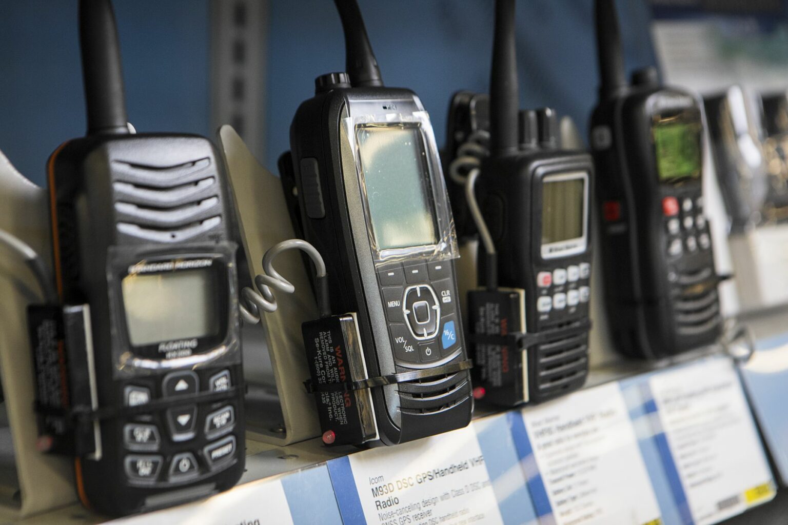 License-free image - Several VHF handheld radios are displayed upright on a shelf in a store. Each radio has a different design and control layout. The shelf below features price tags and descriptions for the radios.