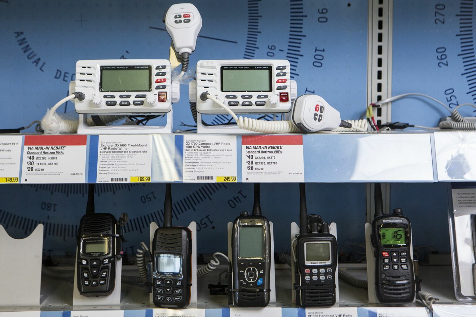 License-free image - A store shelf displays various two-way radios and communication devices. Two white marine radios with microphones sit on the top shelf, while six handheld radios, varying in size and design, are arranged on the lower shelf. Price tags are visible below each item. VHF and handheld VHF radios on display at a retail outlet.