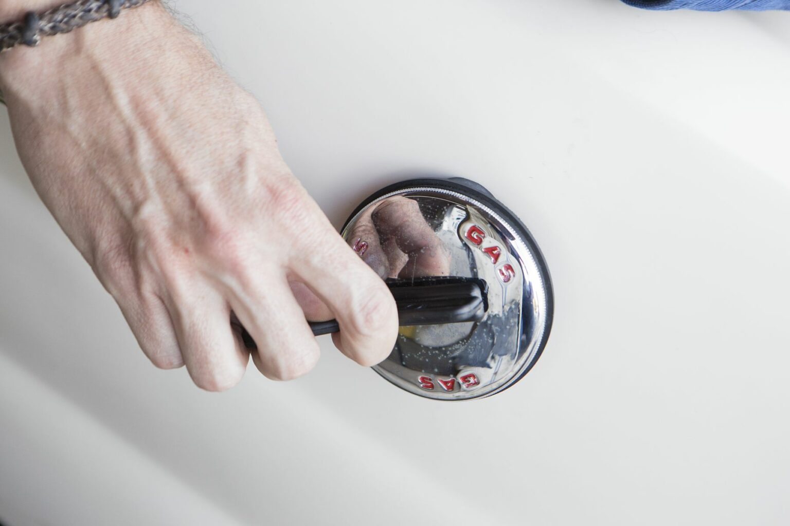 License-free image - A person’s hand gripping and turning a gas cap on a white vehicle. The cap has the word &quot;GAS&quot; written in red letters. The person is wearing a chain bracelet around their wrist. Opening a marine gas cap using a gas cap key.