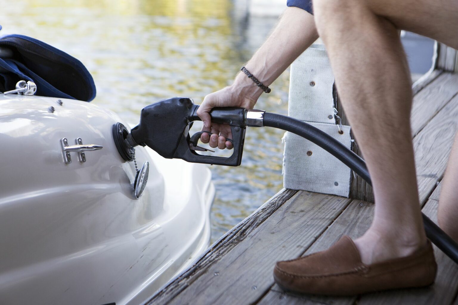 License-free image - Close-up of a person refueling a boat at a dock. The person is holding a fuel pump nozzle and inserting it into the boat&#039;s fuel tank. The dock is wooden, and the water is visible in the background. The person is wearing shorts and brown boat shoes. Refueling a boat at a marina pump.