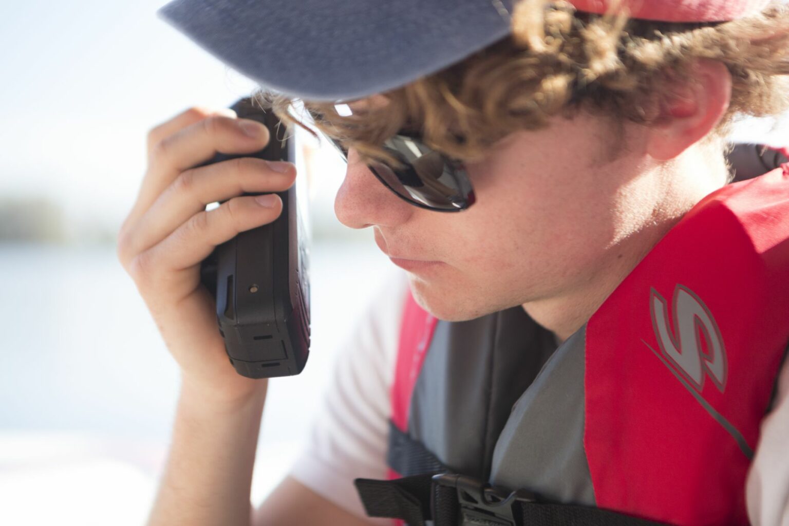 Royalty-free image - A person with curly hair and sunglasses is using a handheld VHF-DSC marine band radio set to channel 16. They are wearing a red and gray life vest and a blue cap while on a boat. The background is a blurred view of the water and sky.