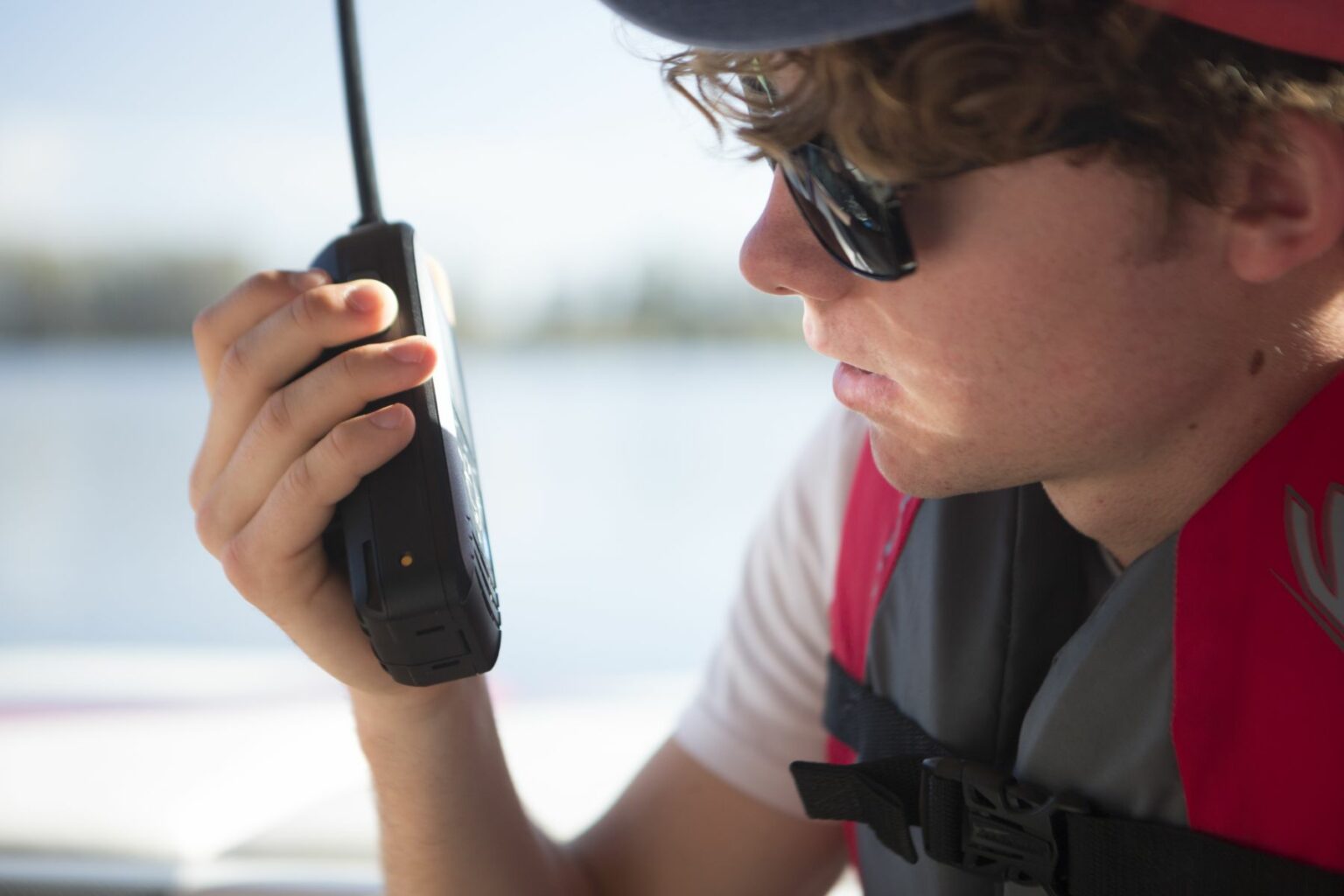 Royalty-free image - A person wearing sunglasses and a life jacket holds a handheld VHF-DSC marine band radio set to channel 16 close to their mouth. The background reveals a blurry view of water and trees, suggesting they are on alert for any maritime communication.