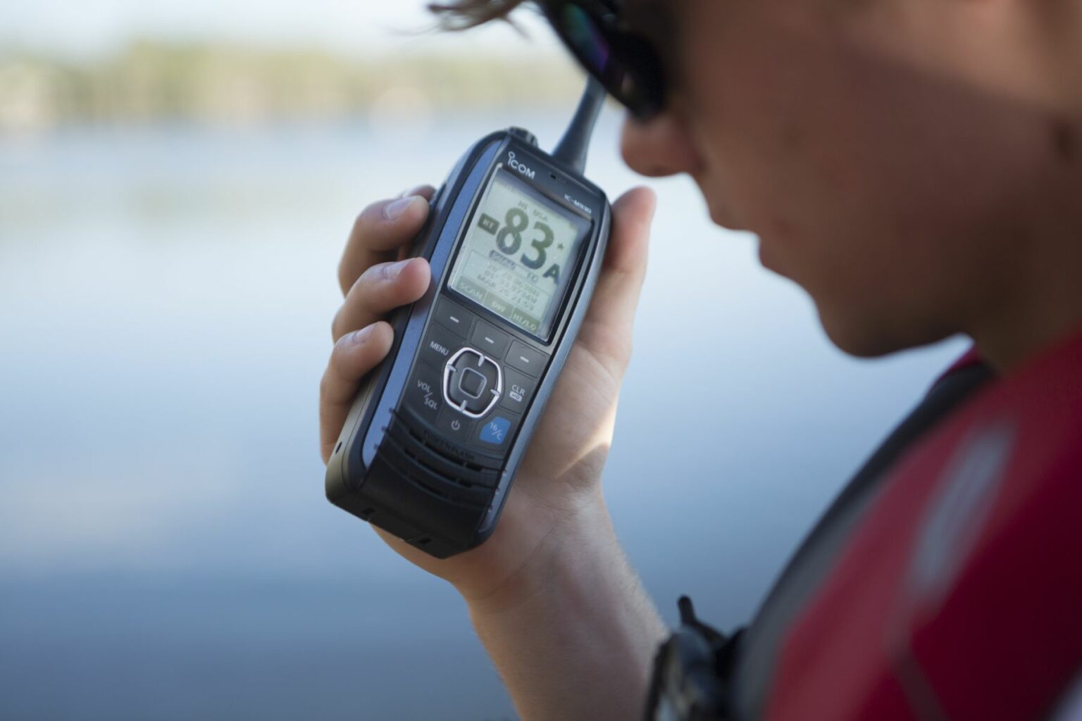 Royalty-free image - A person in a red jacket holds a handheld VHF-DSC marine band radio set to channel 16, intently gazing at the digital screen. The blurred expanse of water in the background hints at an outdoor setting near a lake or river, capturing the tranquility and purpose of the moment.