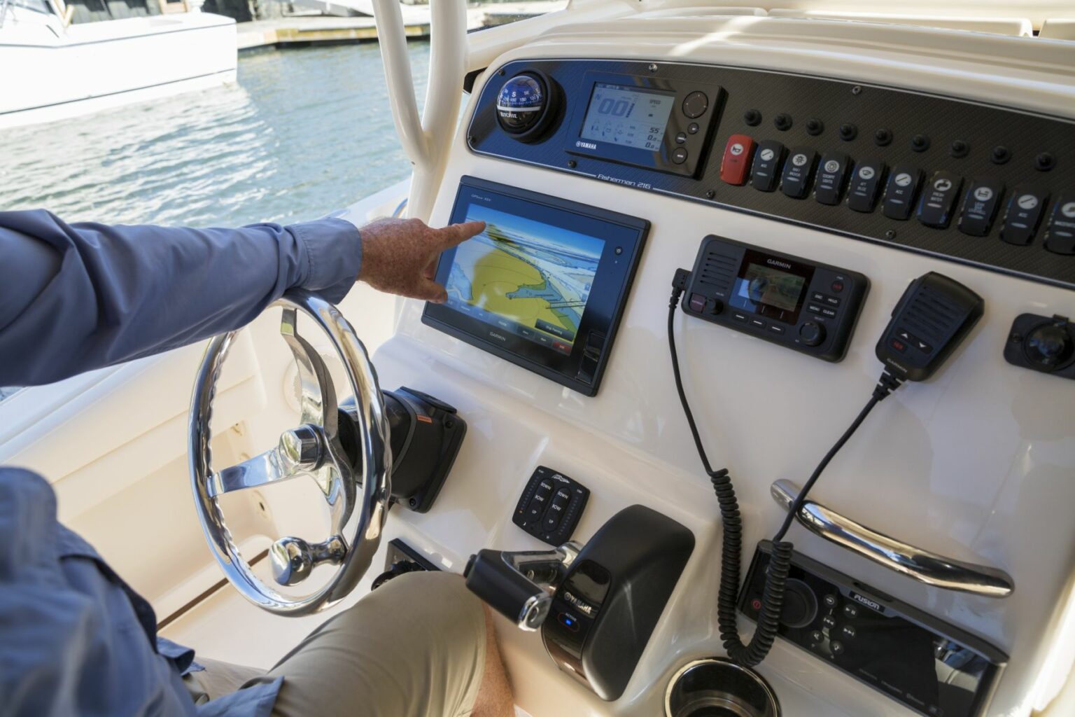 Royalty-free image - Person operating a boat&#039;s control panel, featuring a touchscreen displaying navigation information. Various instruments, knobs, and a wheel are visible. A marina with boats can be seen in the background.