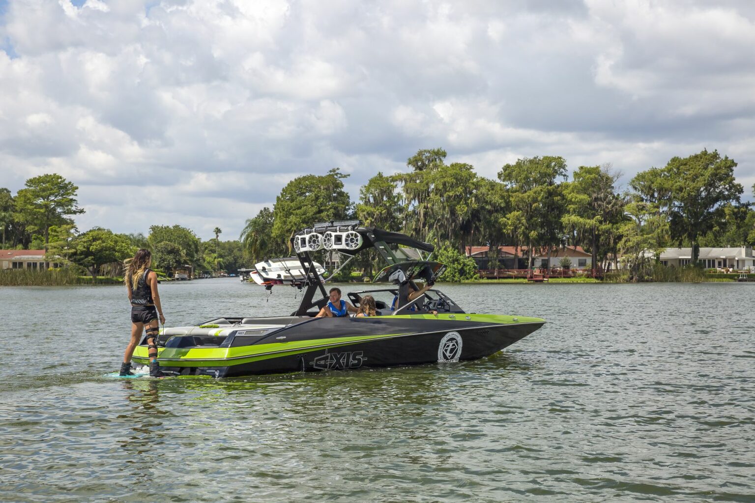 License-free image - A vibrant lime green and black boat with several people on board is floating on a calm lake. One person stands on the edge of the boat while others are seated. The background features trees, houses, and a partly cloudy sky. Wakeboarding behind a wakeboard boat on Clear Lake in Orlando, Florida.