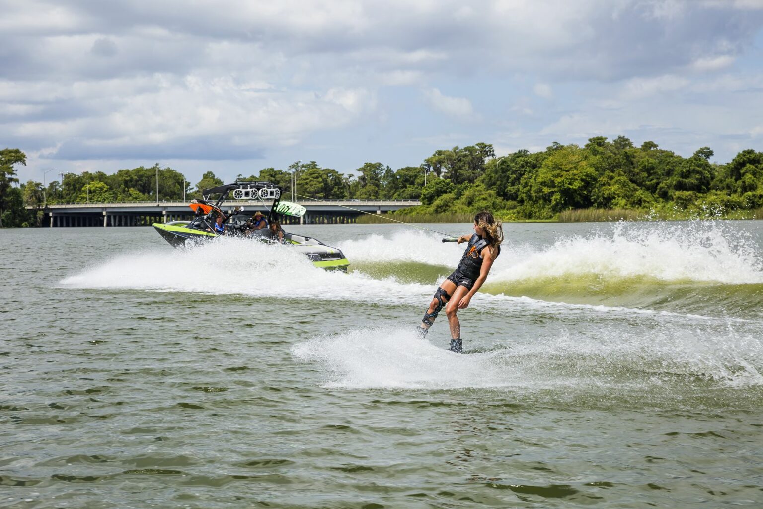 License-free image - A person is wakeboarding on a lake, being pulled by a motorboat with several passengers. The person is performing a trick, creating splashes and waves. The background features a bridge and lush green trees under a partly cloudy sky. Wakeboarding behind a wakeboard boat on Clear Lake in Orlando, Florida.