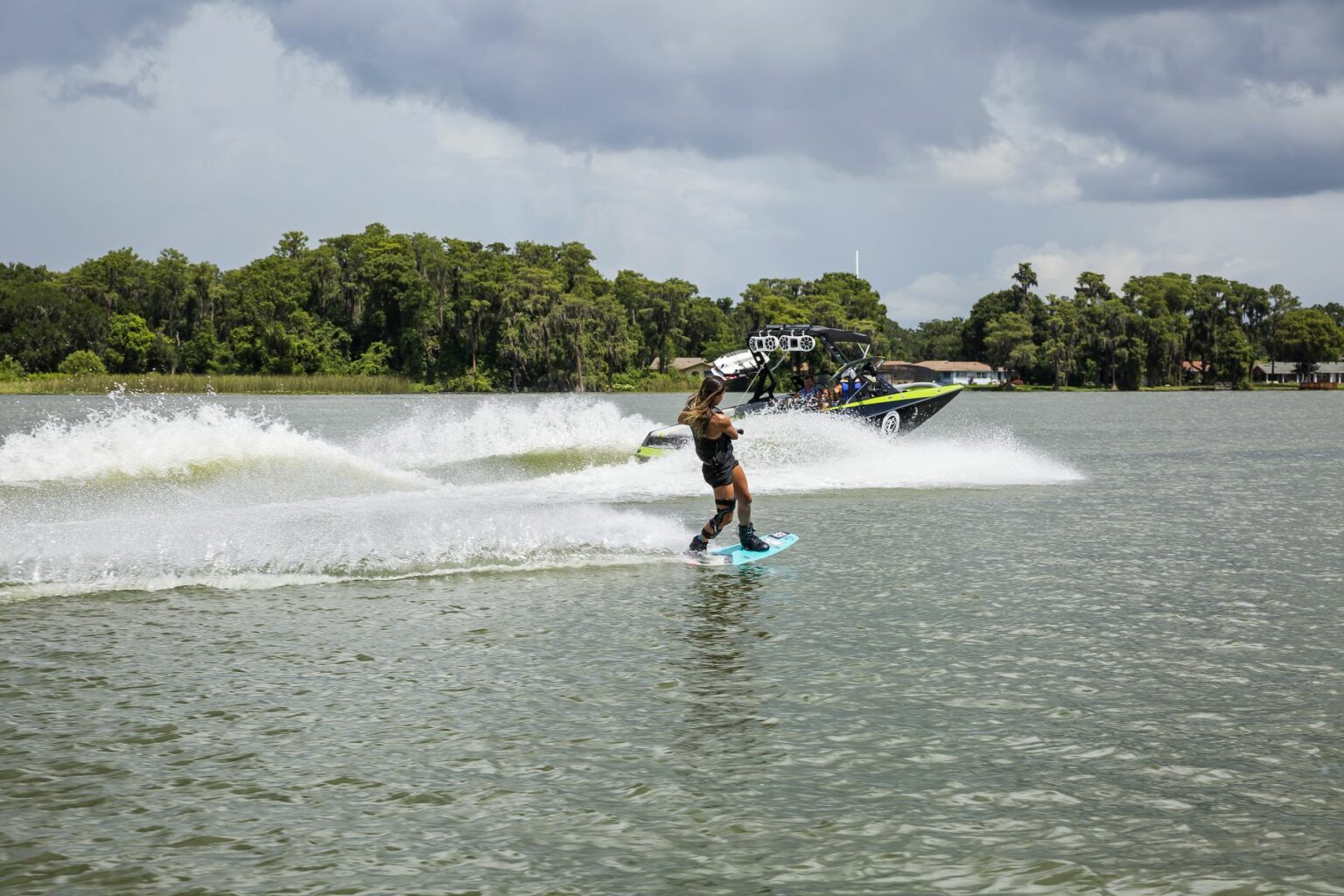 License-free image A person wakeboarding on a lake, being pulled by a motorboat. The wakeboarder is balancing skillfully, creating splashes in the water. The boat is moving quickly, and the background shows a line of trees under a cloudy sky. Wakeboarding behind a wakeboard boat on Clear Lake in Orlando, Florida.