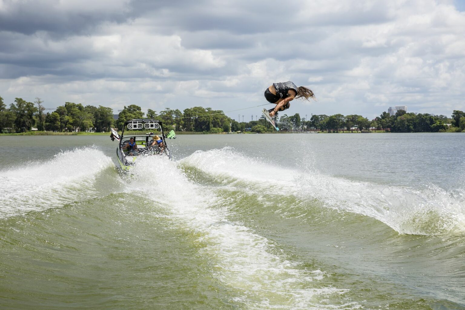 License-free image - A person is mid-air performing a trick on a wakeboard, being towed by a motorboat on a lake. The boat, with several passengers, makes a large wake. The sky is partly cloudy and trees line the distant shore. Wakeboarding behind a wakeboard boat on Clear Lake in Orlando, Florida.