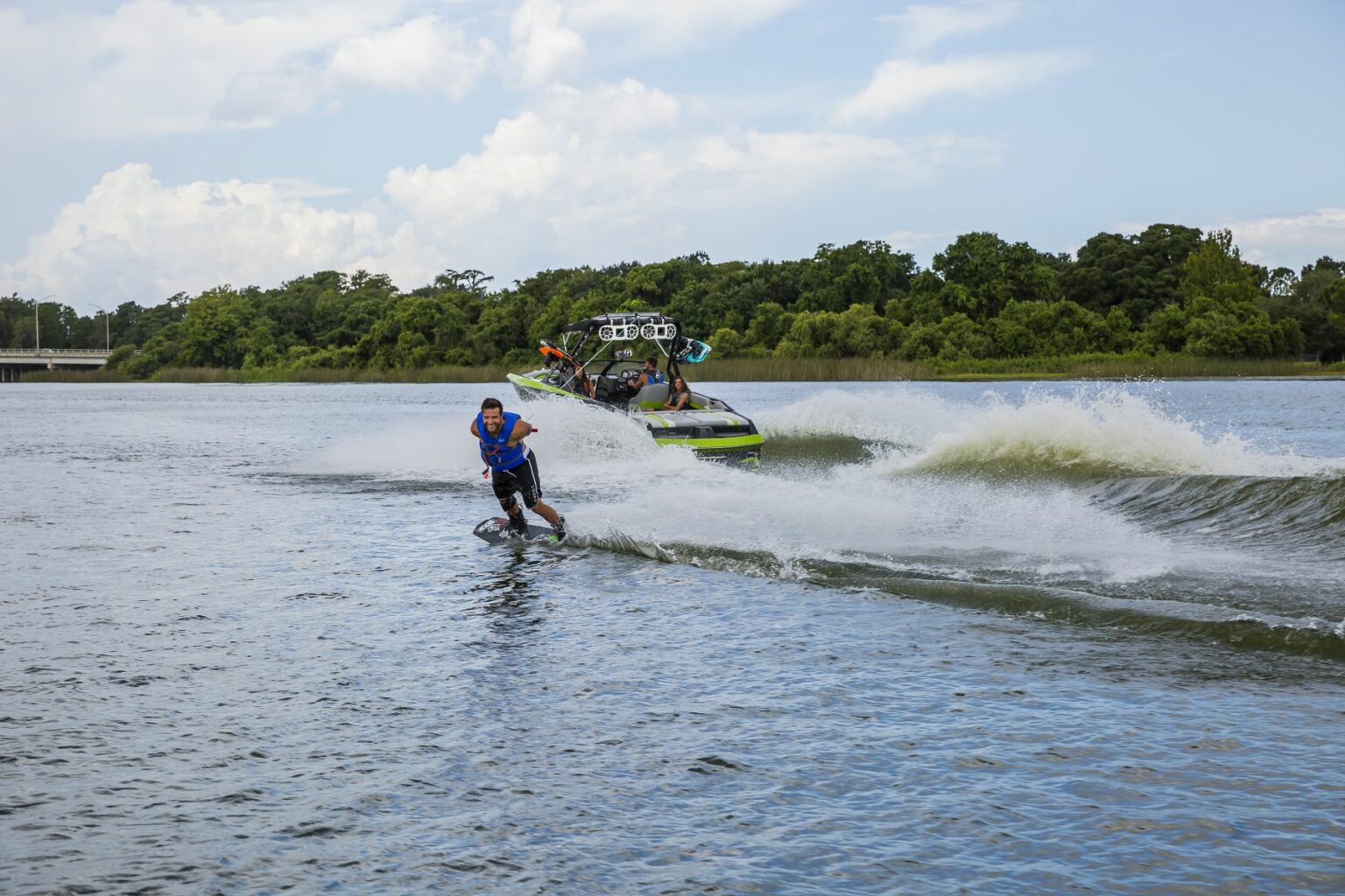 License-free image - A person wakeboarding on a body of water, being pulled by a motorboat with several people on board. The wakeboarder is wearing a blue life jacket and is riding on the water&#039;s surface, creating a splash. The background includes trees and a cloudy sky. Wakeboarding behind a wakeboard boat on Clear Lake in Orlando, Florida.