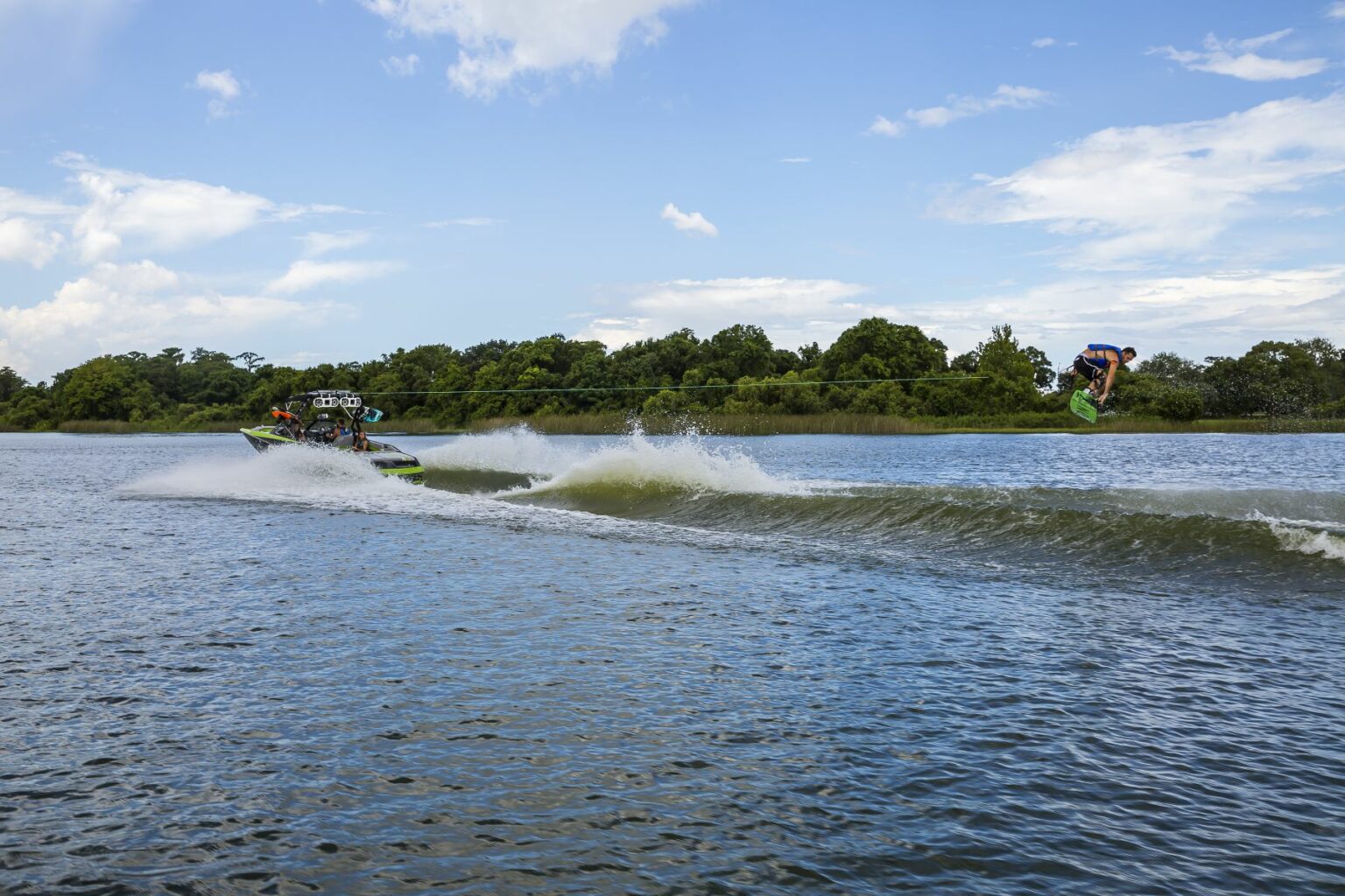 License-free image - A person is performing an aerial trick on a wakeboard, propelled by a green and white speedboat on a large body of water. The wakeboarder is high in the air while holding the board. The background features a line of trees under a partly cloudy sky. Wakeboarding behind a wakeboard boat on Clear Lake in Orlando, Florida.