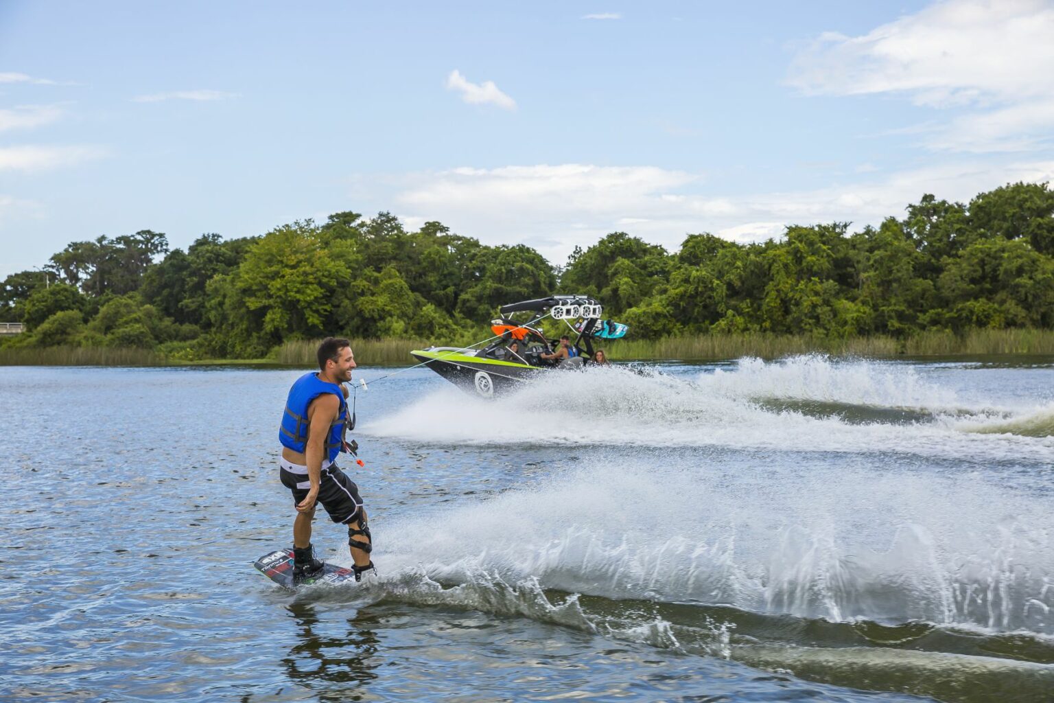 License-free image - A person in a blue life jacket is wakeboarding on a lake behind a motorboat. The boat, with several passengers on board, creates a wake in the water. The background features lush green trees and a partly cloudy sky. Wakeboarding behind a wakeboard boat on Clear Lake in Orlando, Florida.