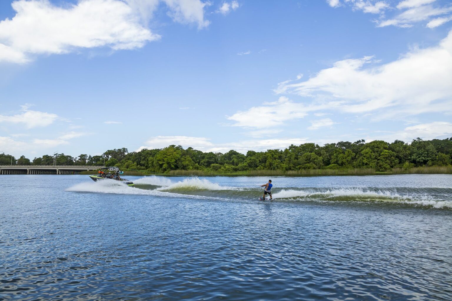 License-free image A person is wakeboarding on a large body of water, being towed by a motorboat. The boat leaves a trail of white water and waves behind it. Lush green trees line the distant shoreline under a blue sky with scattered clouds. Wakeboarding behind a wakeboard boat on Clear Lake in Orlando, Florida.