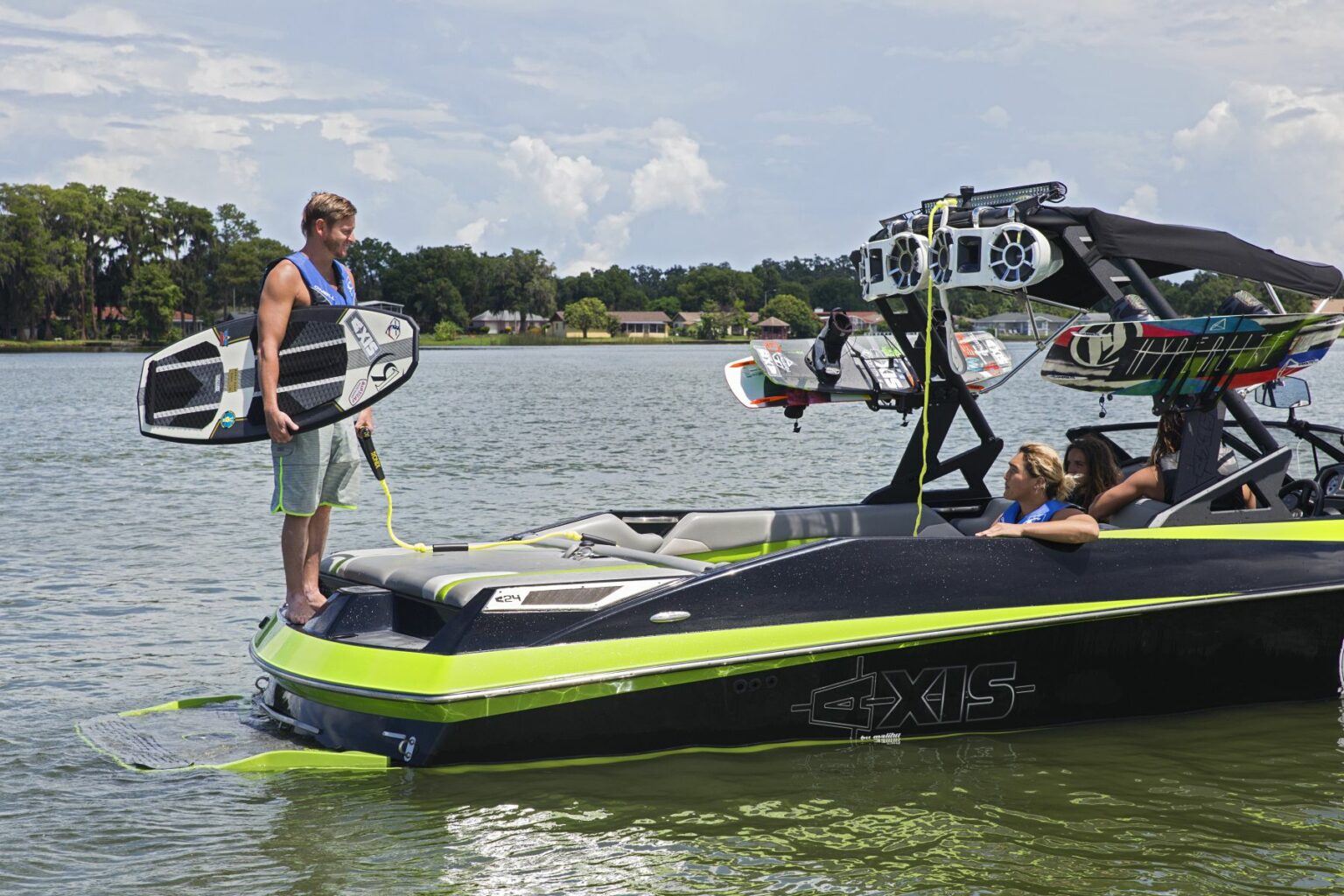 License-free image - A person stands on the swim platform of a green and black Axis boat holding a wakesurf board. Another person is sitting inside the boat, and additional boards are mounted on the boat&#039;s tower. The scene takes place on a calm lake with trees and houses in the background. Wakesurfing behind a wakeboard boat on Clear Lake in Orlando, Florida.