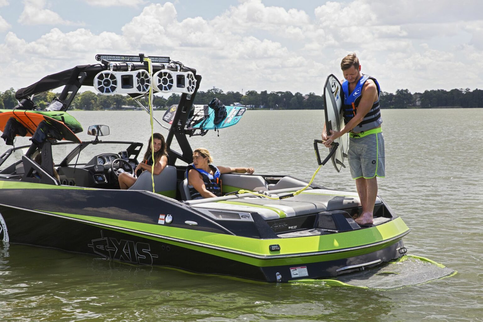 License-free image - A man stands on the back of a green and black Axis boat holding a wakeboard, preparing to get into the water. Two women are sitting in the boat, wearing life jackets and smiling. The boat is equipped with speakers, and a calm lake surrounds them.