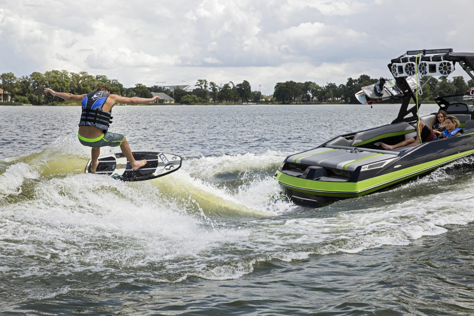 License-free image - A person in a life vest and shorts is wakeboarding behind a black and green boat driven by another person. The wakeboarder is mid-air performing a trick, with surrounding trees and a partly cloudy sky in the background. Wakesurfing behind a wakeboard boat on Clear Lake in Orlando, Florida.