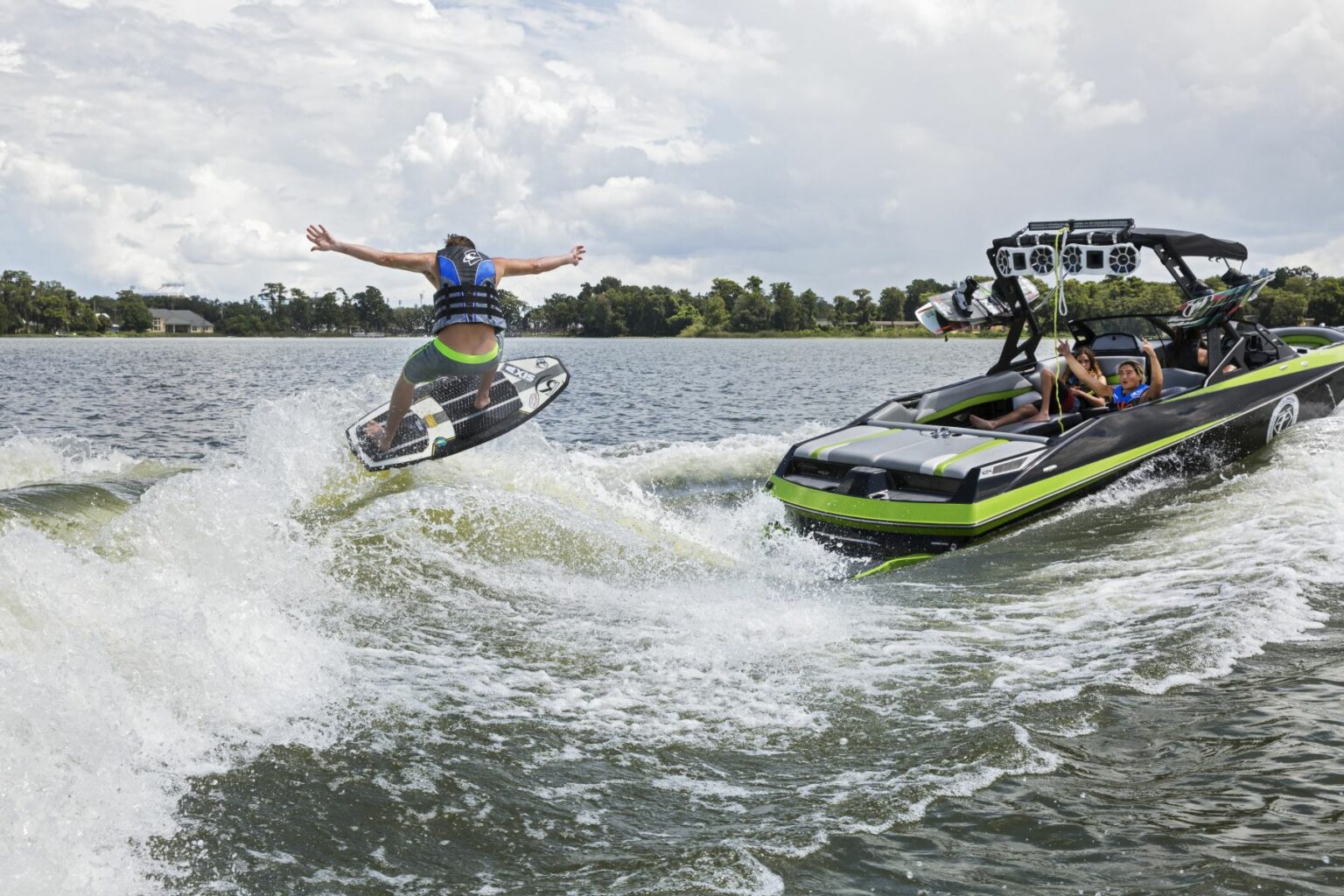 Royalty-free image - A person on a wakeboard performs a jump over a wave while being towed by a green and black boat. Another person in the boat is watching, and there are trees and cloudy skies in the background. Wakesurfing behind a wakeboard boat on Clear Lake in Orlando, Florida.