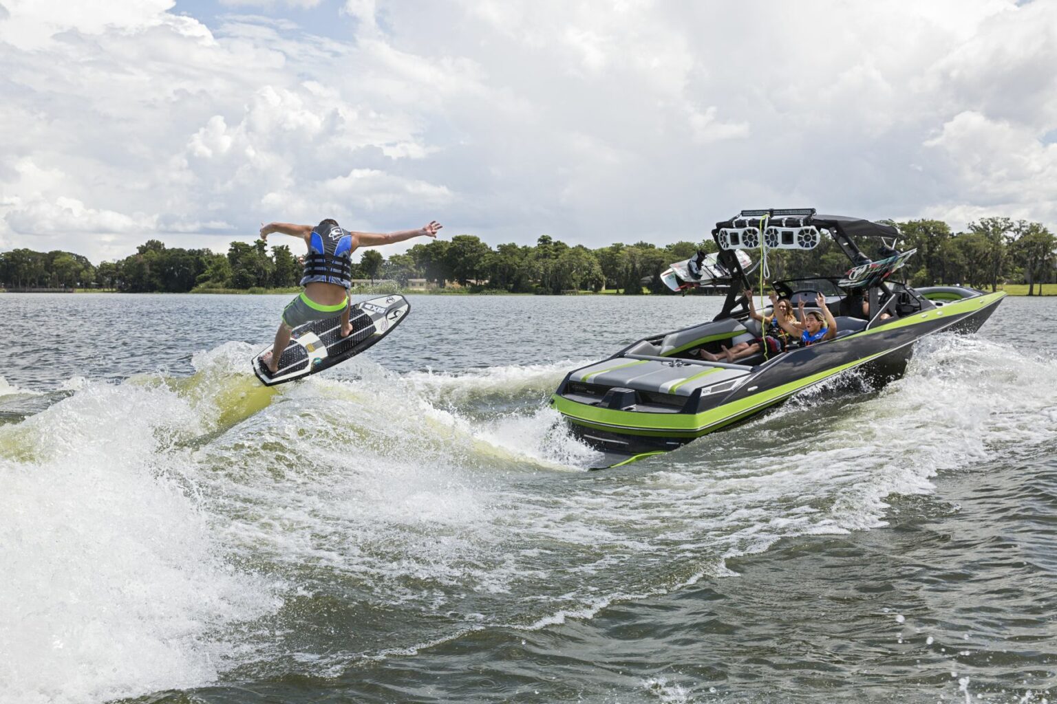 License-free image - A person wearing a life jacket jumps high on a wakeboard over the wake created by a speedboat with several passengers on a sunny day. The boat is sleek and black with green accents, cruising on a calm lake with trees and cloudy skies in the background. Wakesurfing behind a wakeboard boat on Clear Lake in Orlando, Florida.