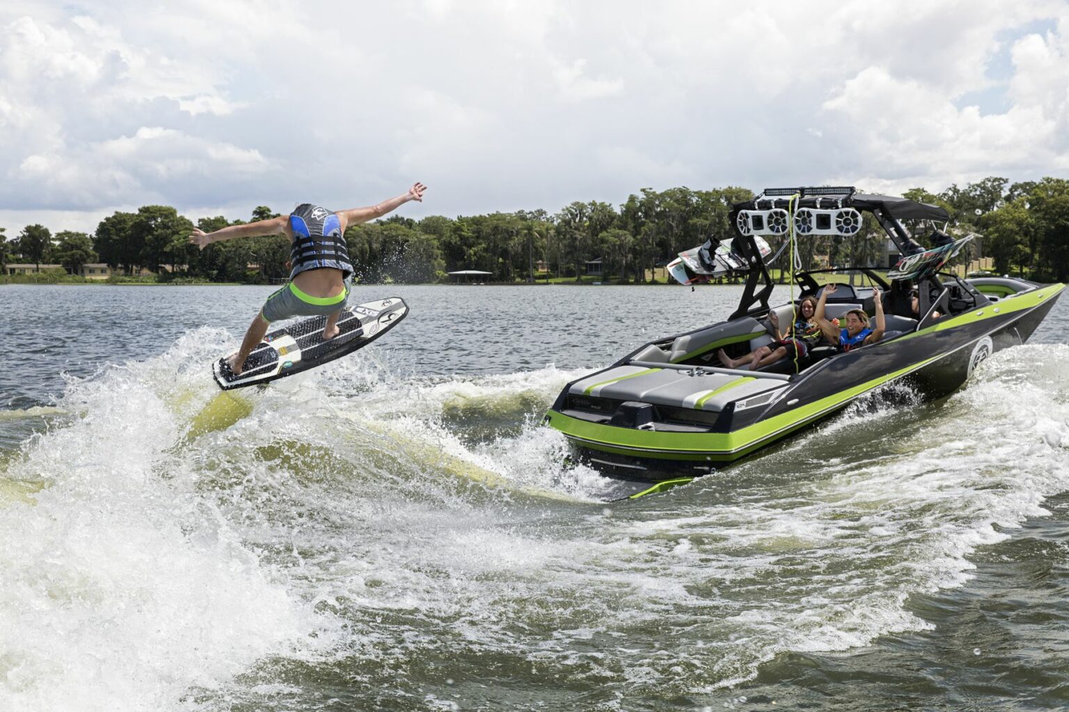 License-free image A person is wakeboarding behind a black and green boat on a sunny day. The wakeboarder is mid-jump, wearing a life jacket and helmet. Two people in the boat are observing the wakeboarder, and the boat is creating waves in the water. Trees and a cloudy sky are in the background. Wakesurfing behind a wakeboard boat on Clear Lake in Orlando, Florida.