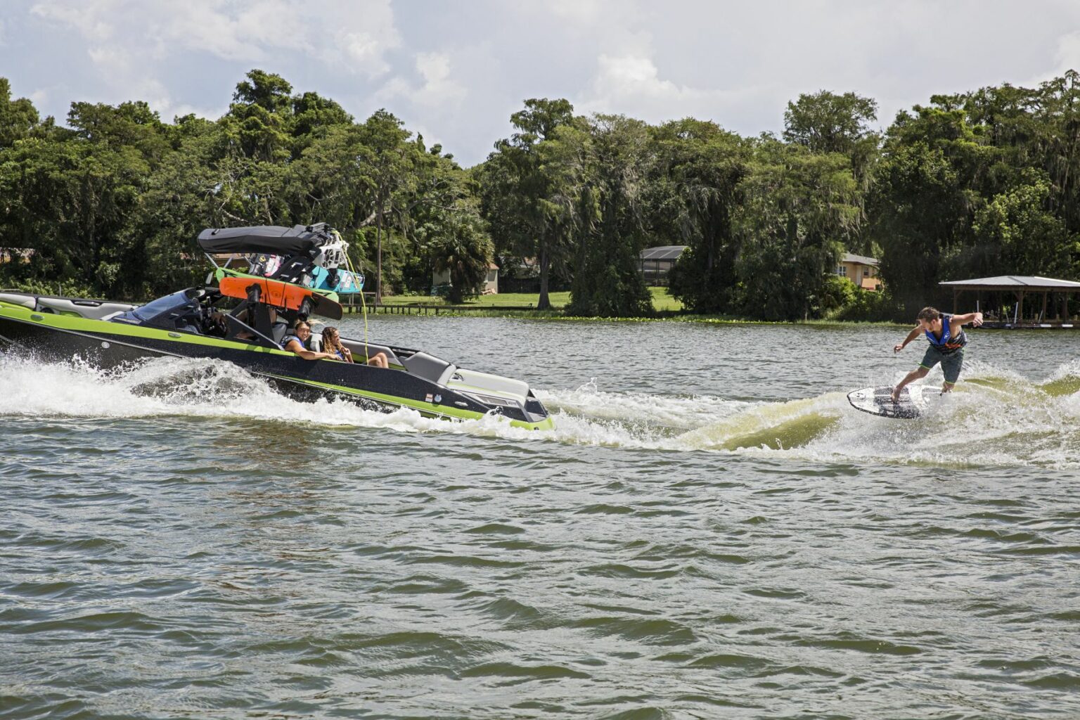 License-free image - A person is wakeboarding behind a motorboat on a lake surrounded by trees. The boat has people inside watching the wakeboarder, and the wakeboarder is riding the wake created by the boat. The sky is partly cloudy. Wakesurfing behind a wakeboard boat on Clear Lake in Orlando, Florida.