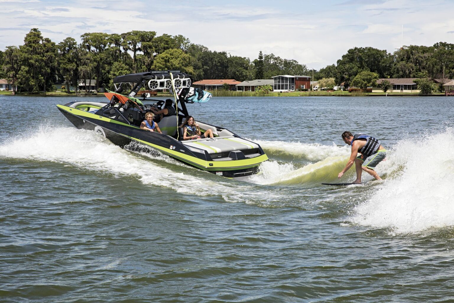 License-free image - A person wearing a life vest rides a wakesurf board on the water near a black and green wakeboard boat. The boat has passengers onboard, and the lake shoreline with trees and buildings is visible in the background under a partly cloudy sky. Wakesurfing behind a wakeboard boat on Clear Lake in Orlando, Florida.