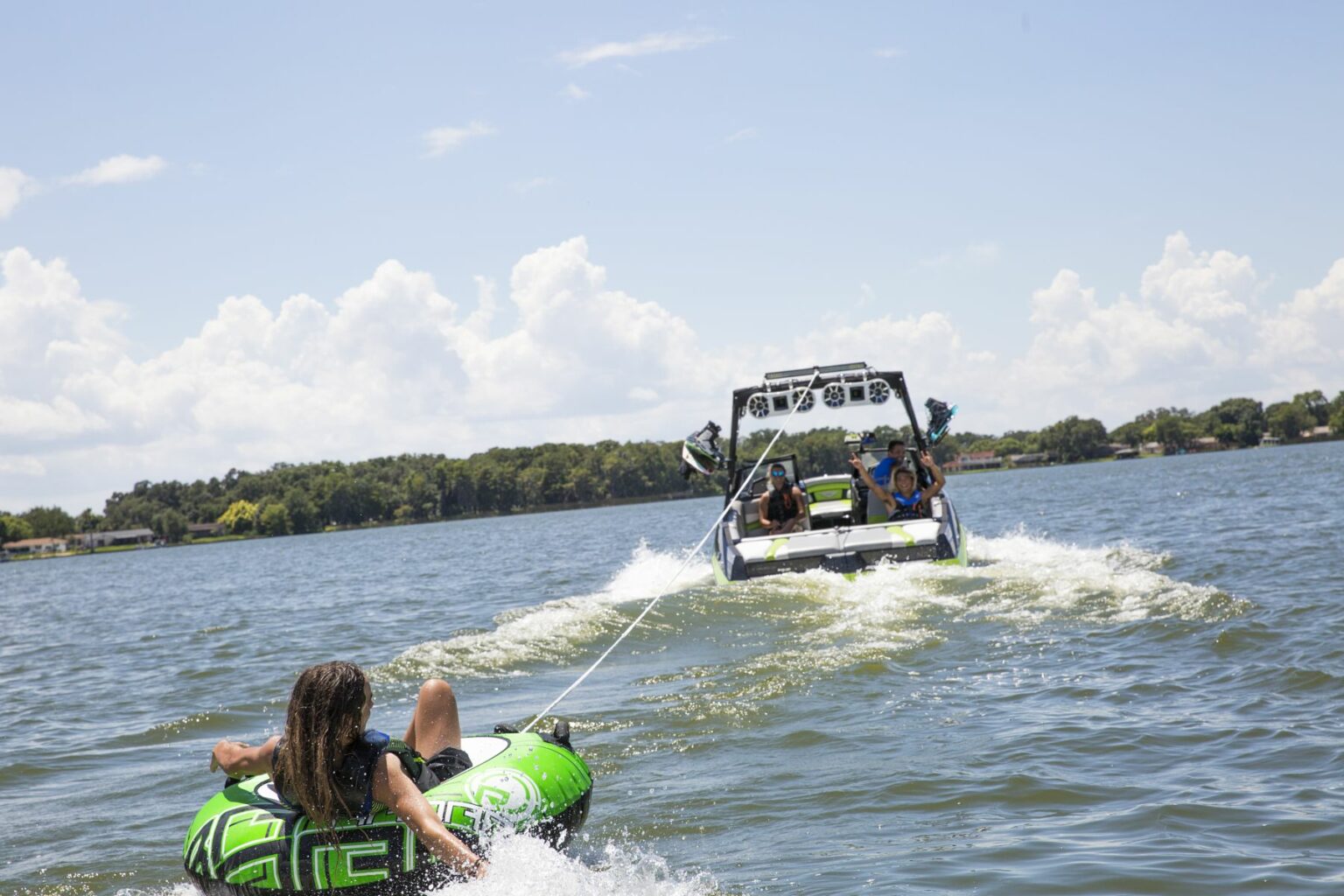 License-free image - A person rides an inflatable tube towed by a speedboat on a lake. The rider, holding onto handles on the green tube, is skimming over the water. The speedboat, with three people on board, creates a white wake under a partly cloudy sky. Forested shoreline is in the distance. Tubing behind a wakeboard boat on Clear Lake in Orlando, Florida.