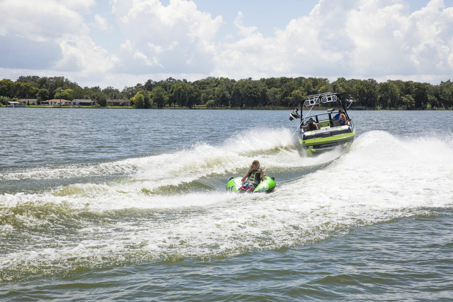 License-free image - A boat pulls a person on an inflatable tube across a lake on a sunny day. The water splashes around the tube, and the boat creates waves in its wake. There are trees and houses in the background under a partly cloudy sky. Tubing behind a wakeboard boat on Clear Lake in Orlando, Florida.