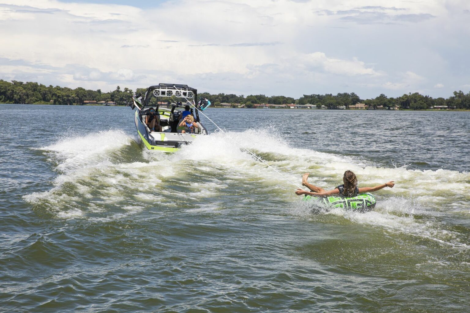 License-free image - A person being towed on an inflatable tube by a speedboat on a sunny day. The boat creates waves as it moves through the water, with the person holding onto the tube and appearing to enjoy the ride. Trees and houses are visible along the shoreline in the background. Tubing behind a wakeboard boat on Clear Lake in Orlando, Florida.