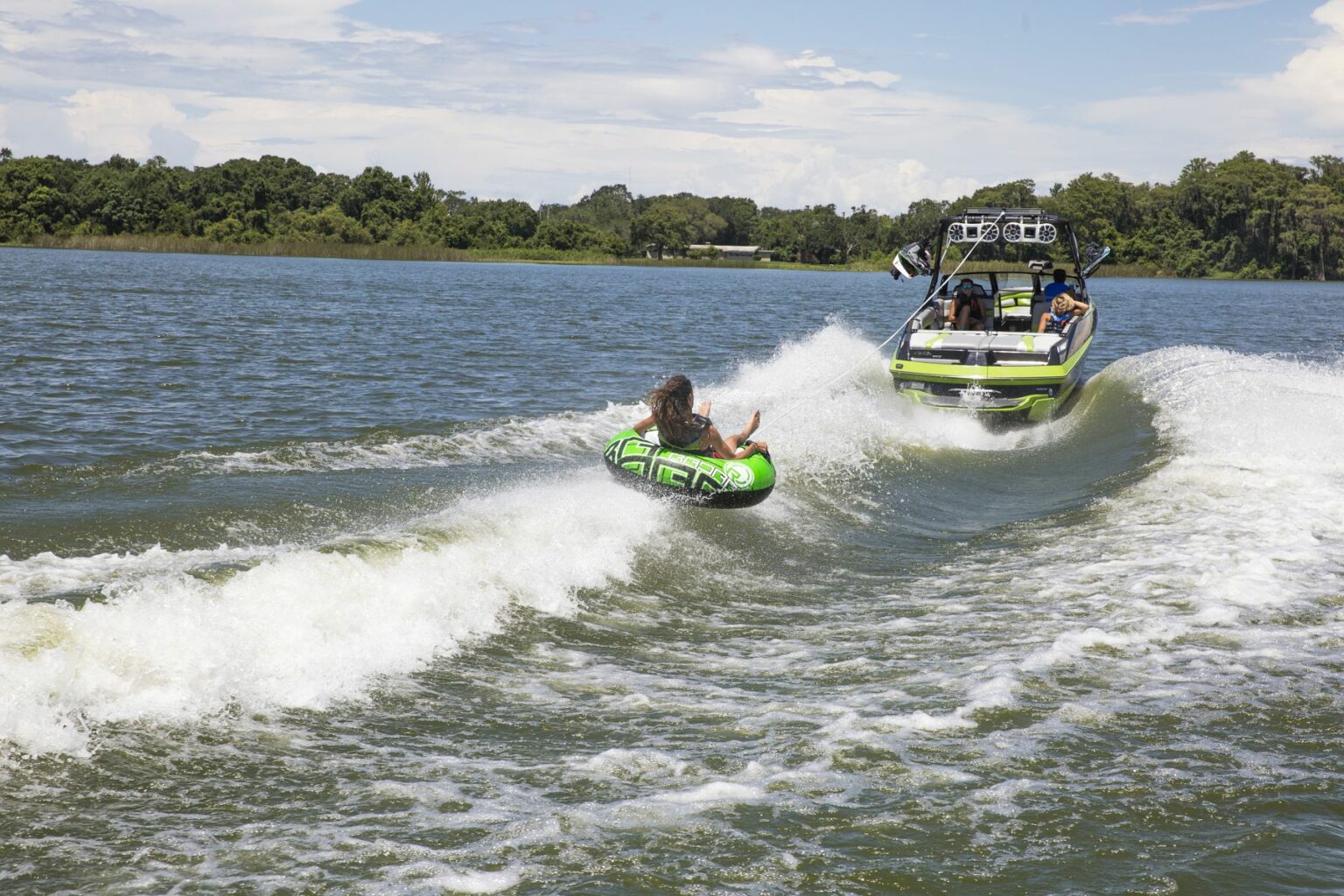 License-free image - Two people sit in a green inflatable tube being towed by a motorboat on a lake. The boat creates white waves, and the tube riders hold on while the scene is surrounded by greenery and a blue sky with scattered clouds.